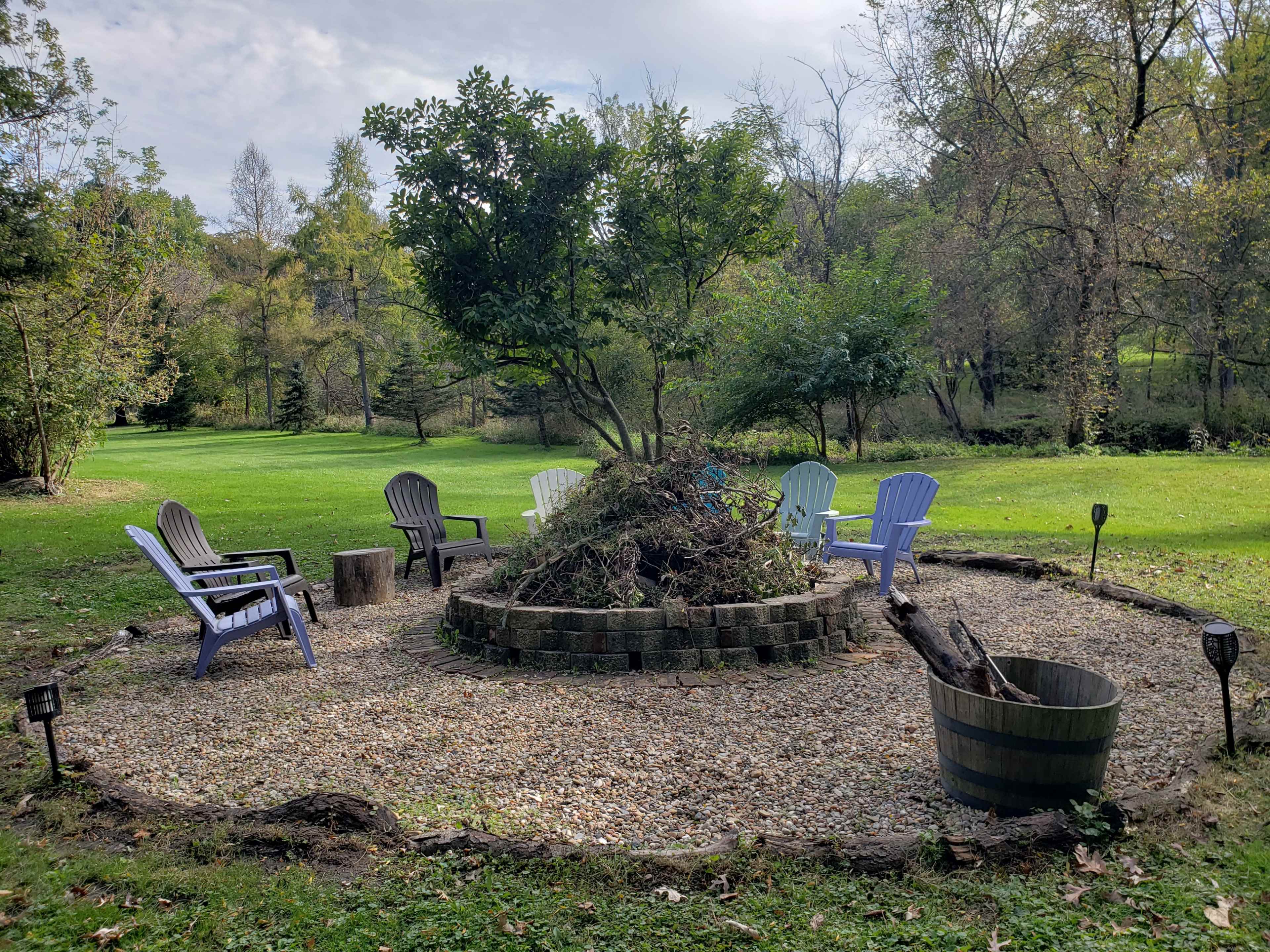 A circular fire pit area surrounded by chairs and a pile of garden debris, situated on a grassy landscape.