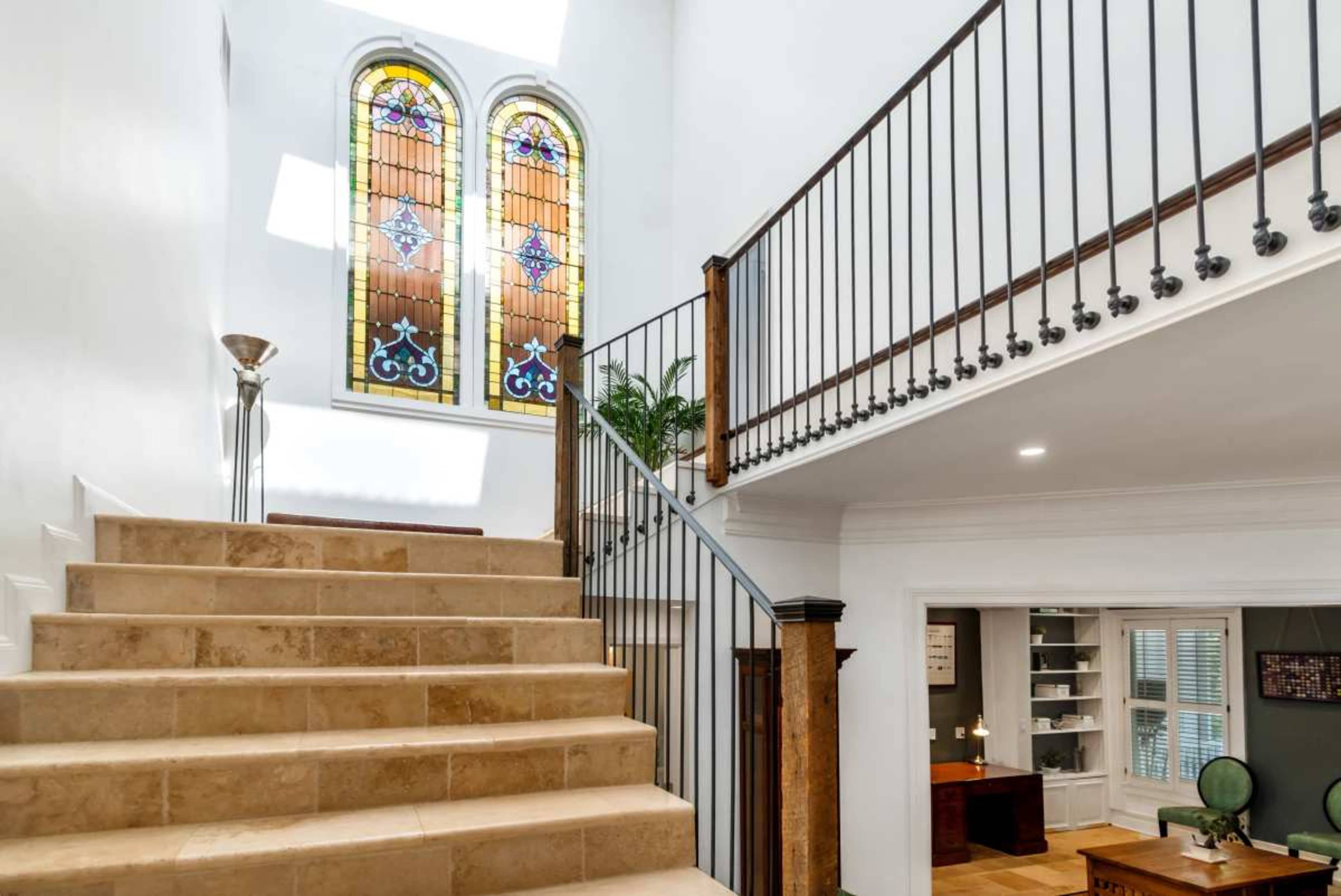 The image shows a staircase with a wrought iron railing leading to a second level, featuring large stained glass windows above that illuminate the space.