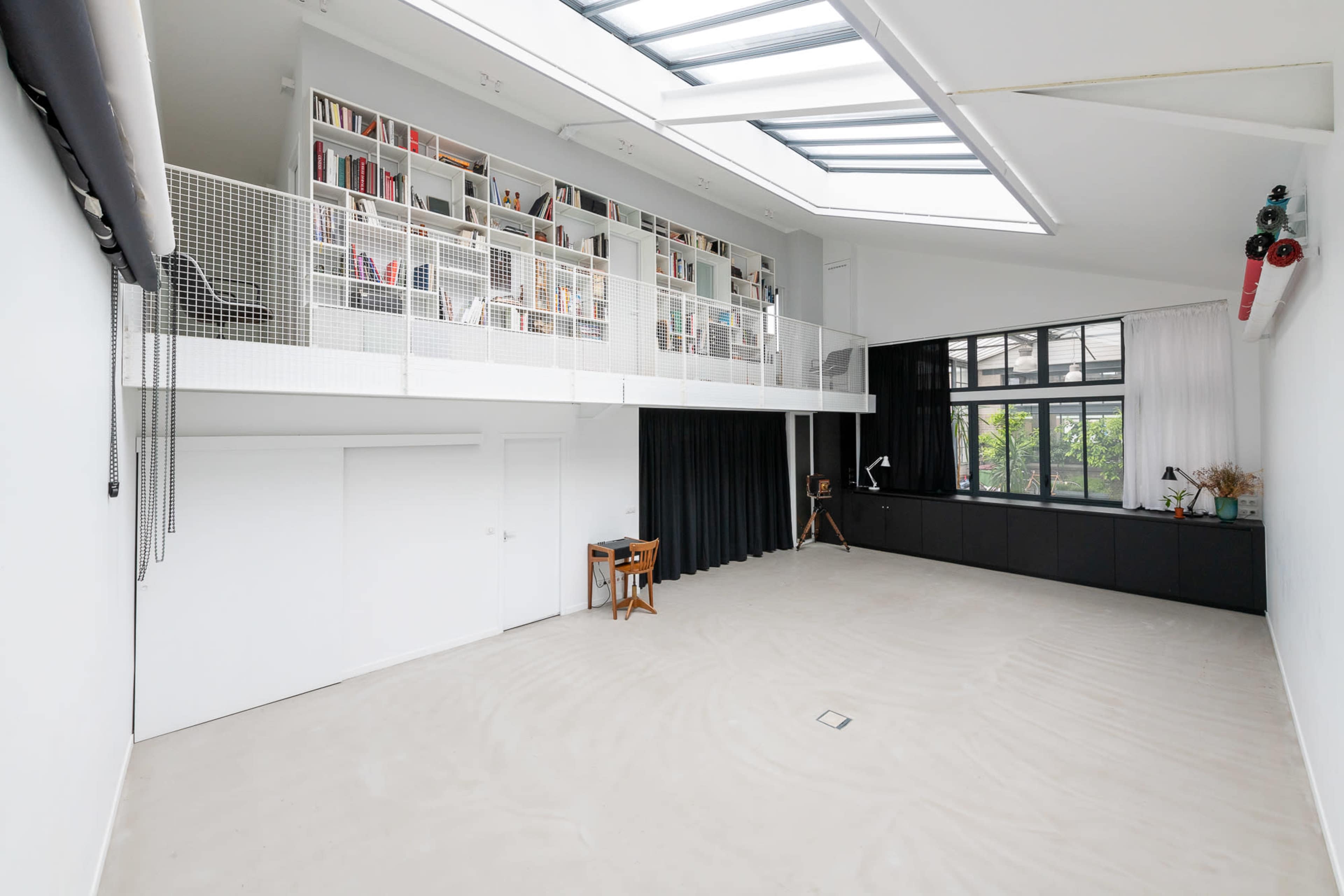 A spacious, modern interior with a high ceiling, a loft area lined with bookshelves, and large windows allowing natural light to enter.