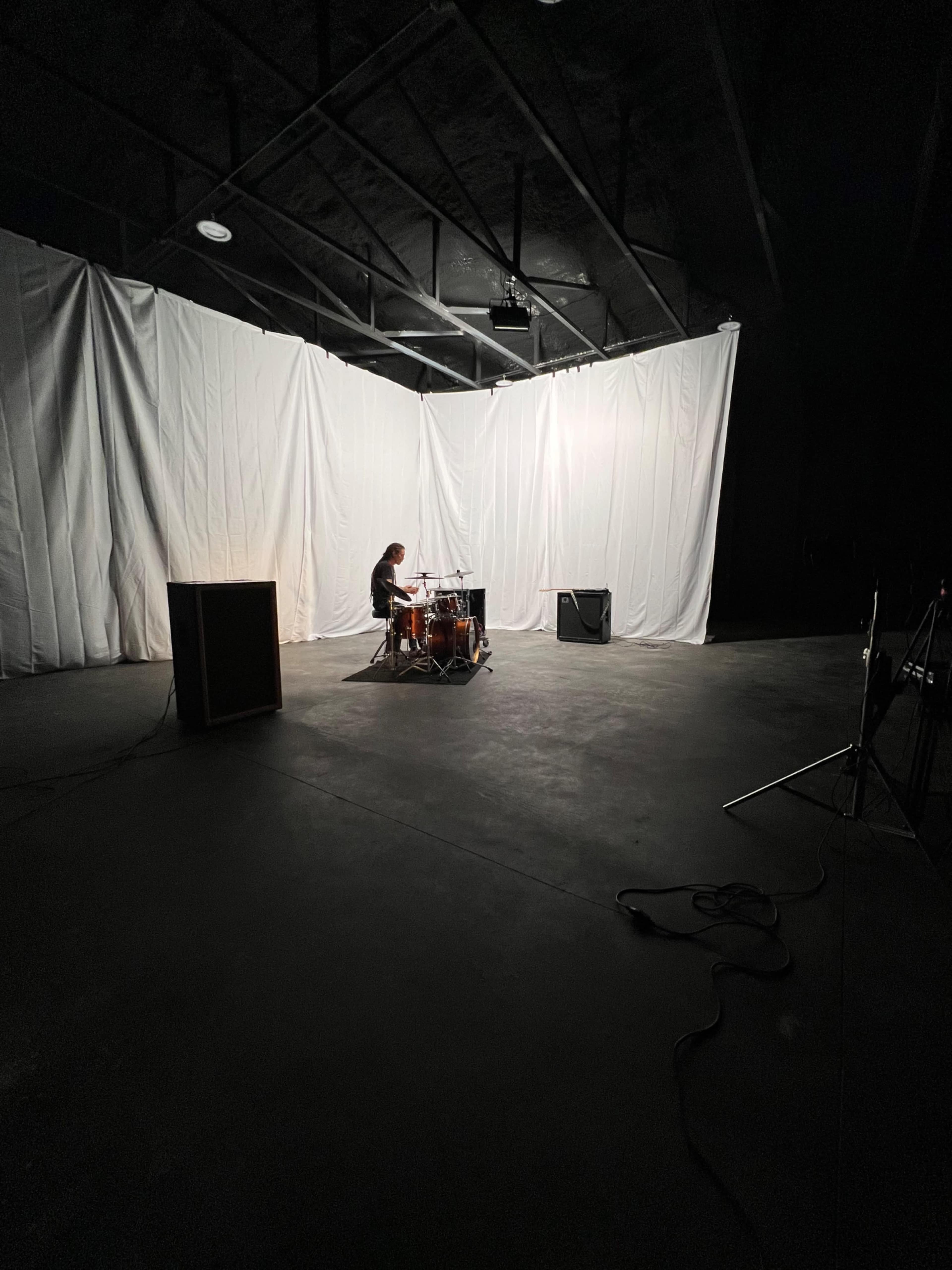 A drummer is seated at a drum set in a dimly lit space with white draping in the background.
