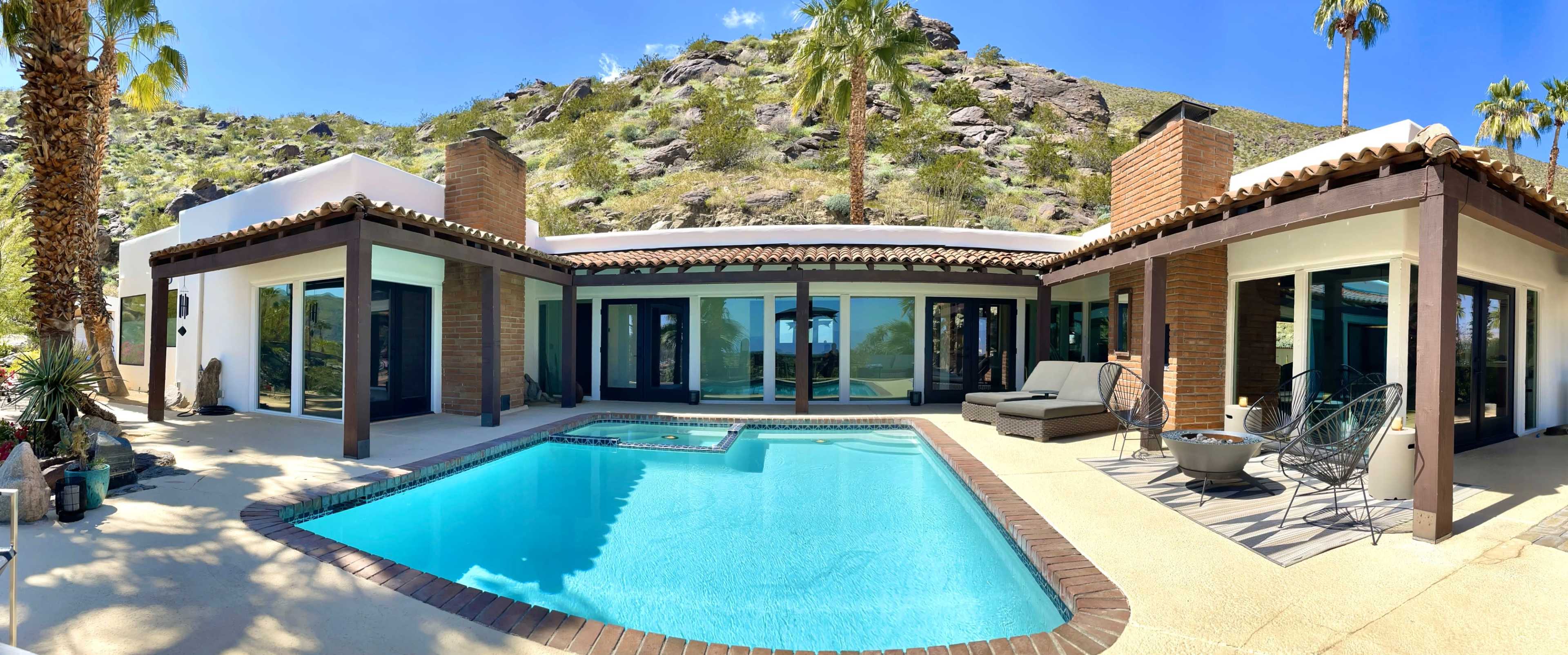 A modern home with a pool is surrounded by palm trees and a hillside, featuring large glass windows and a spacious patio area.
