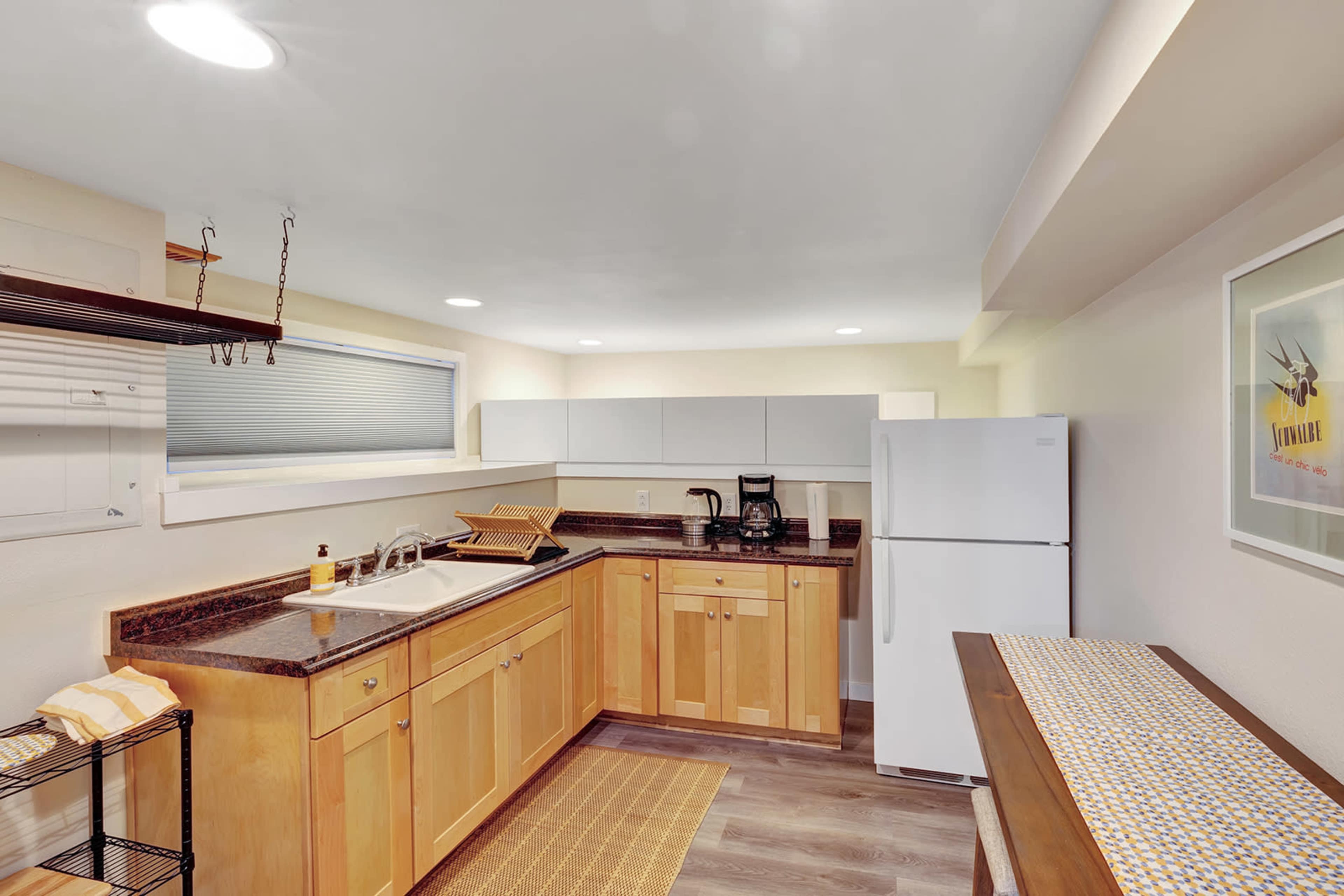 The image shows a modern kitchen with wooden cabinetry, a stainless steel refrigerator, and a countertop work area under bright ceiling lights.