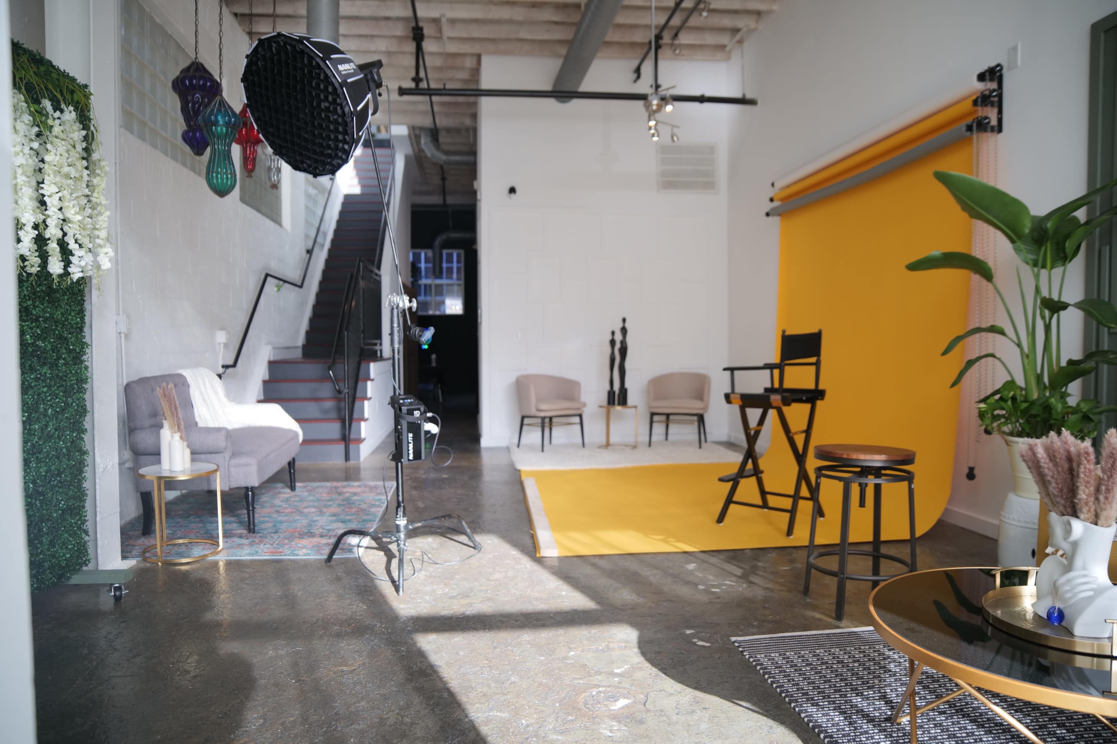 A brightly lit studio space featuring a yellow backdrop, two chairs, a sitting area, and various photography equipment.