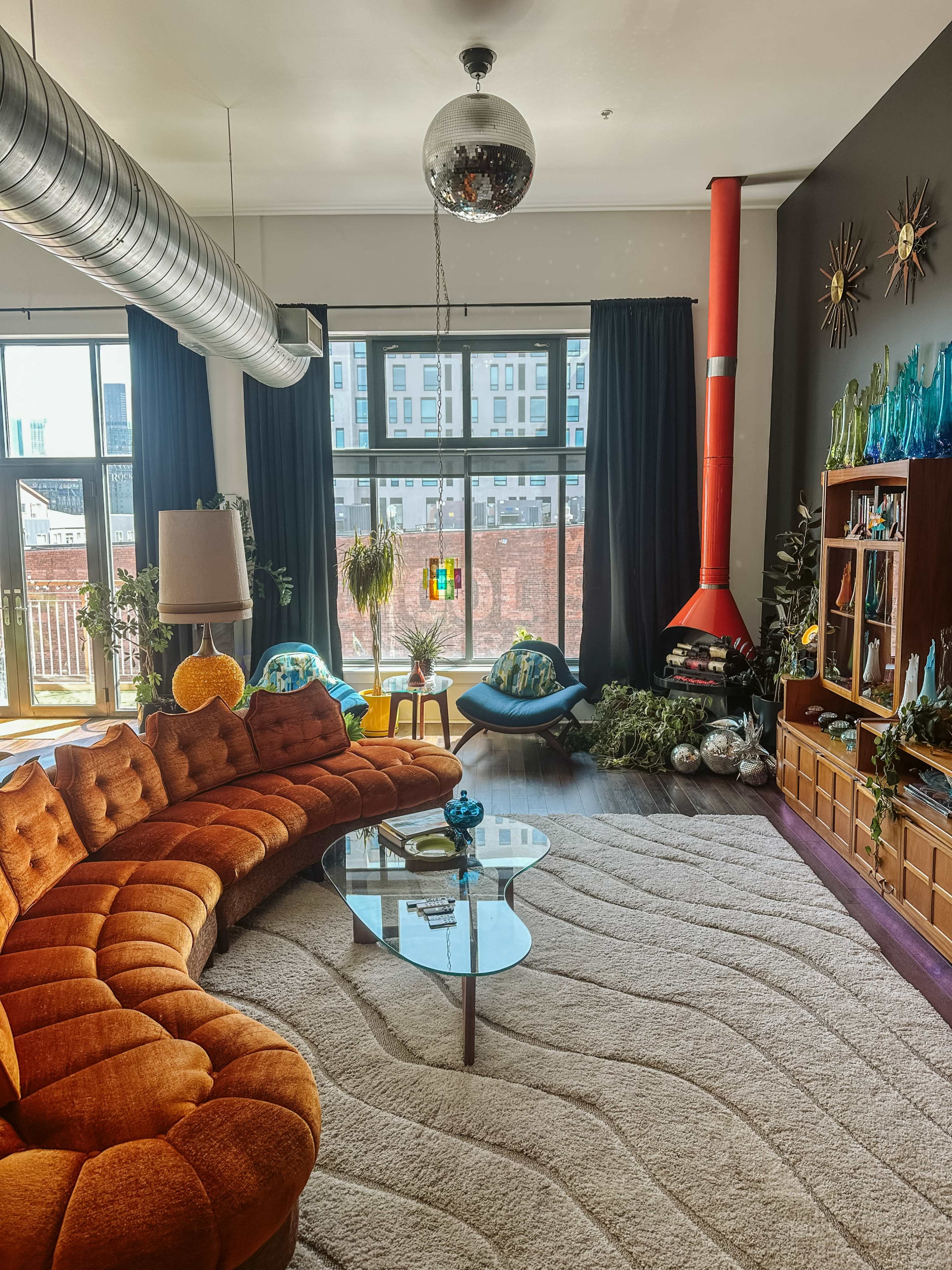 The living room features a curved orange sofa, a glass coffee table, and large windows with a view of the city, complemented by modern decor and plants.
