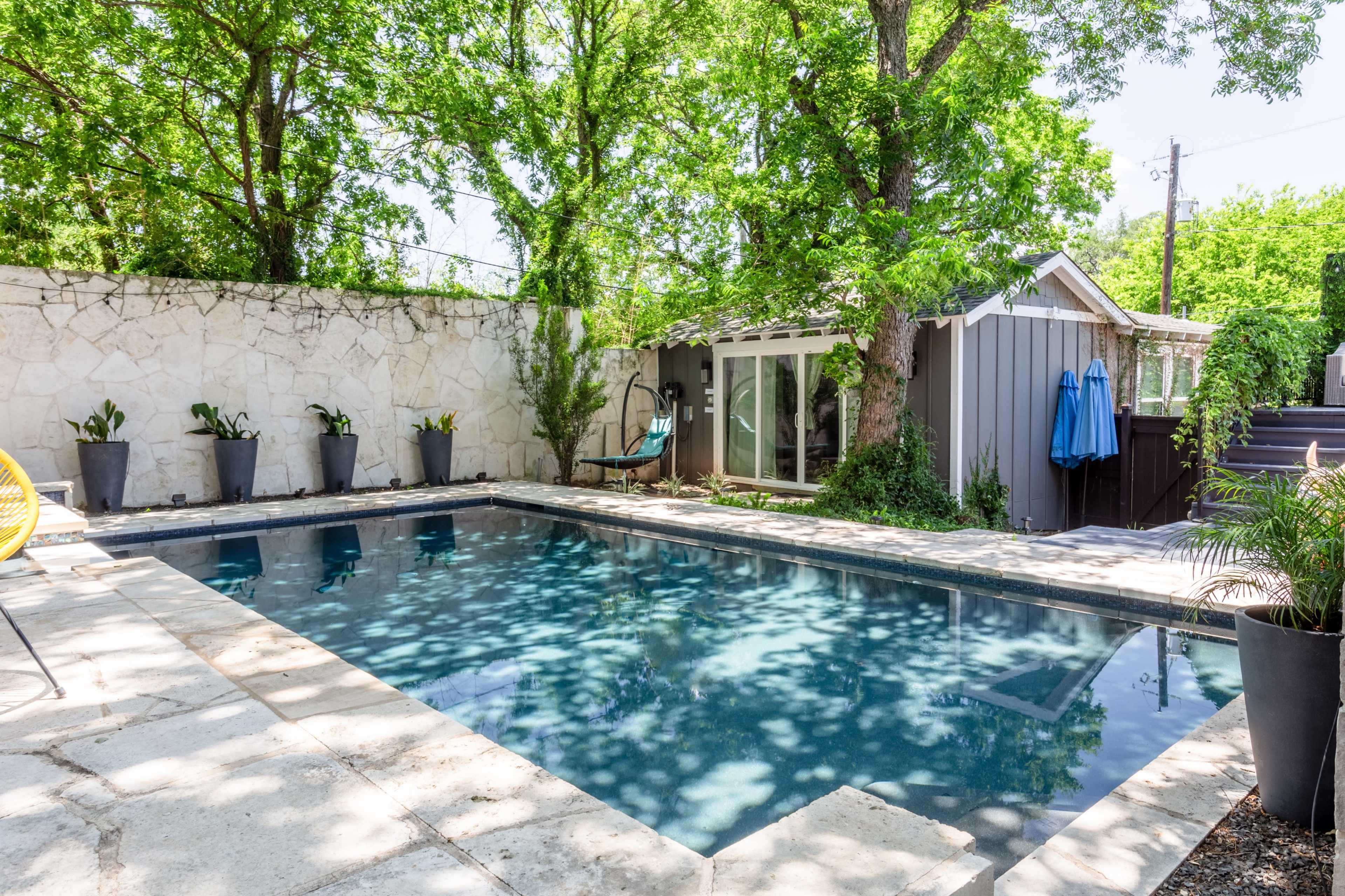 The image shows a rectangular swimming pool surrounded by trees, a stone wall, and a small building with a large window.