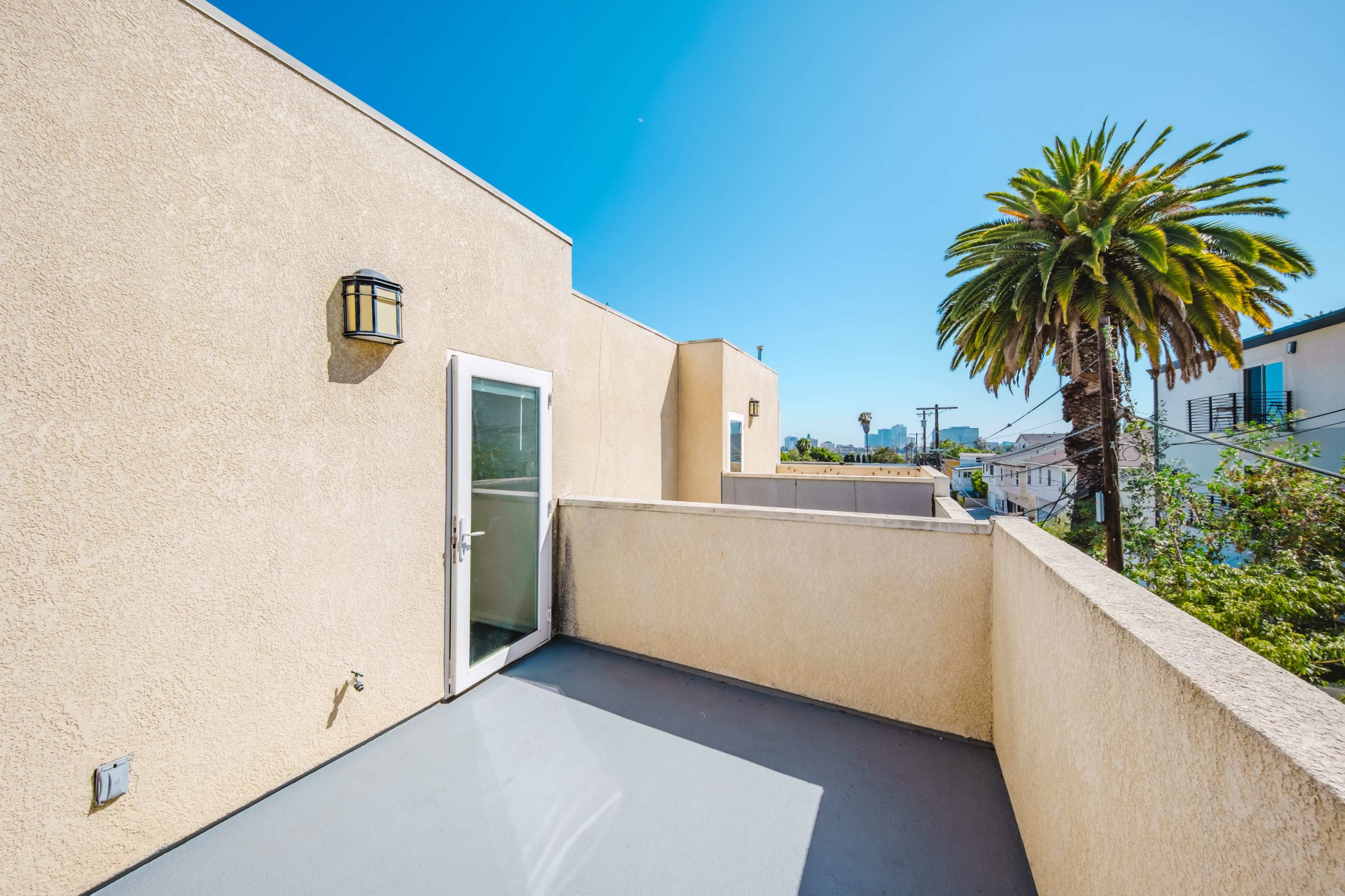 A balcony with a door leading indoors, surrounded by light-colored walls and featuring a palm tree and buildings in the background under a clear blue sky.