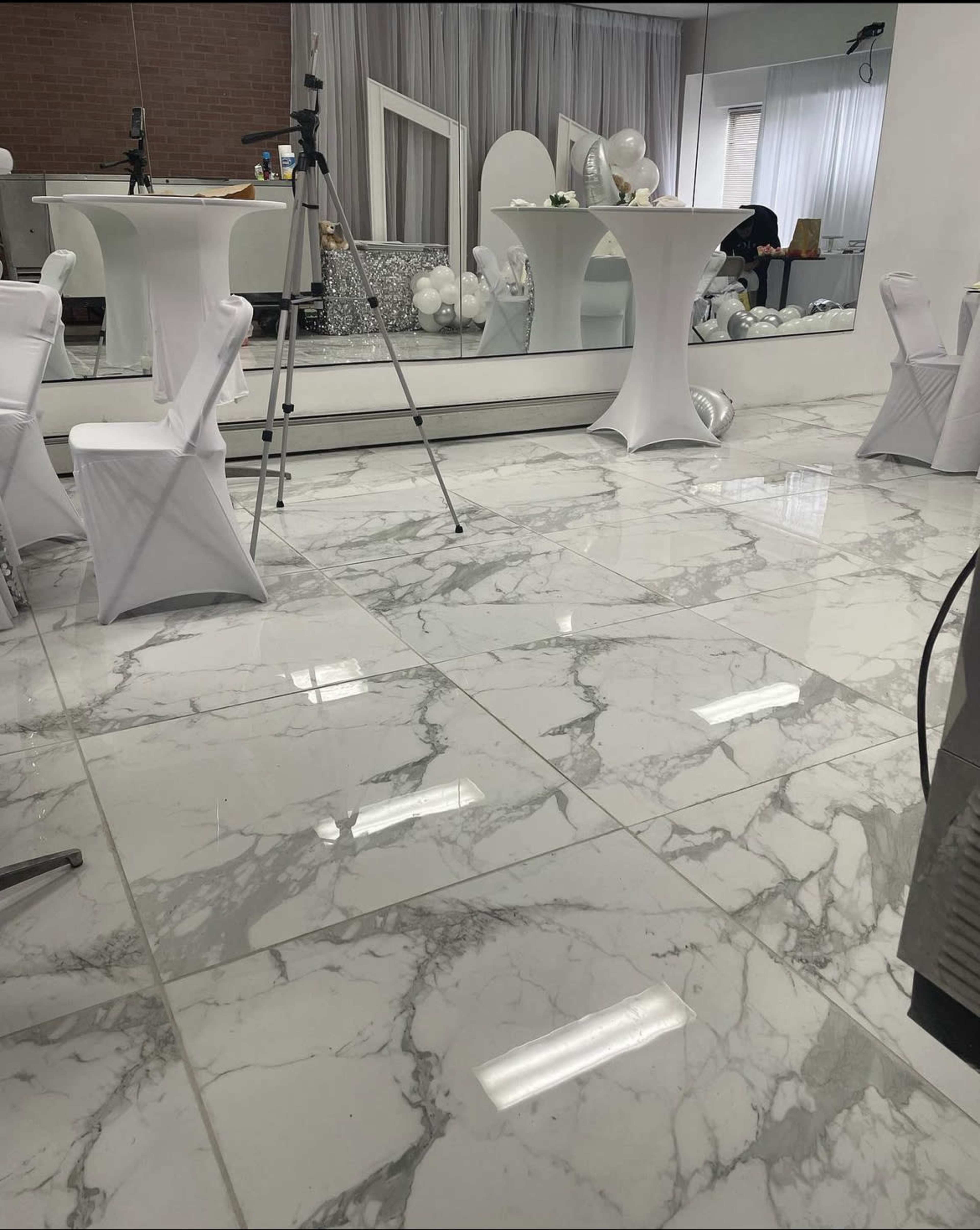 The image shows a spacious indoor event space with white marble flooring, decorated tables, and a backdrop for photography.