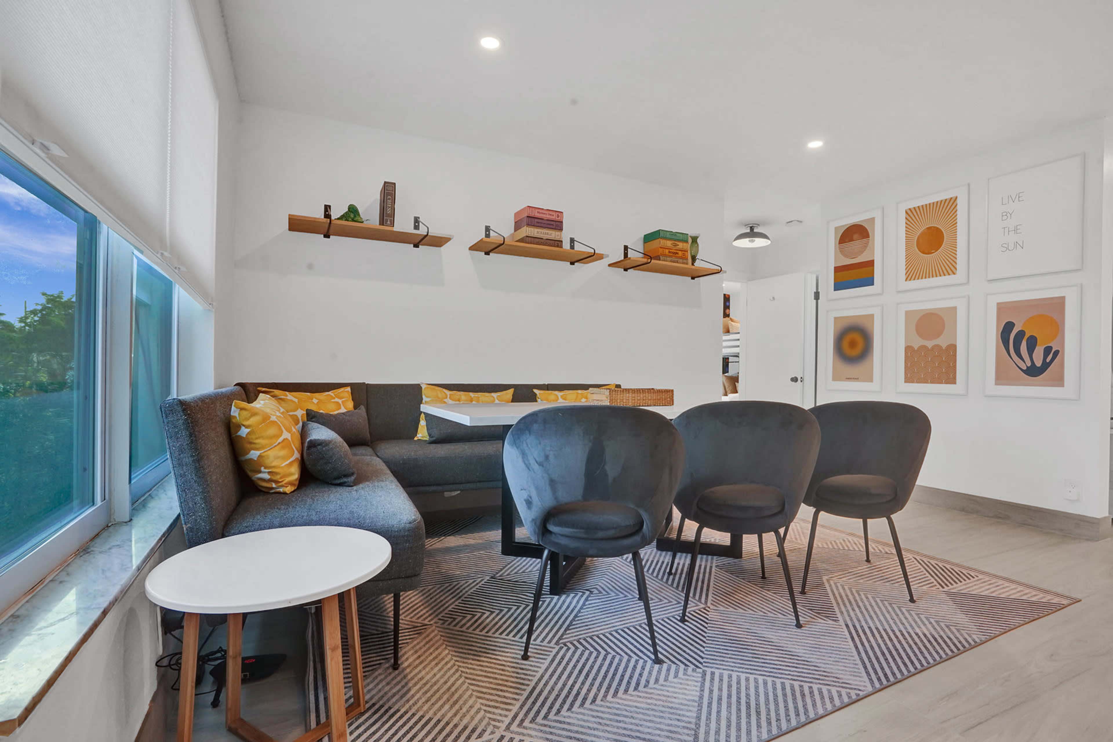 A modern living area with a gray sofa, four black chairs arranged around a patterned rug, and colorful artwork on the wall.
