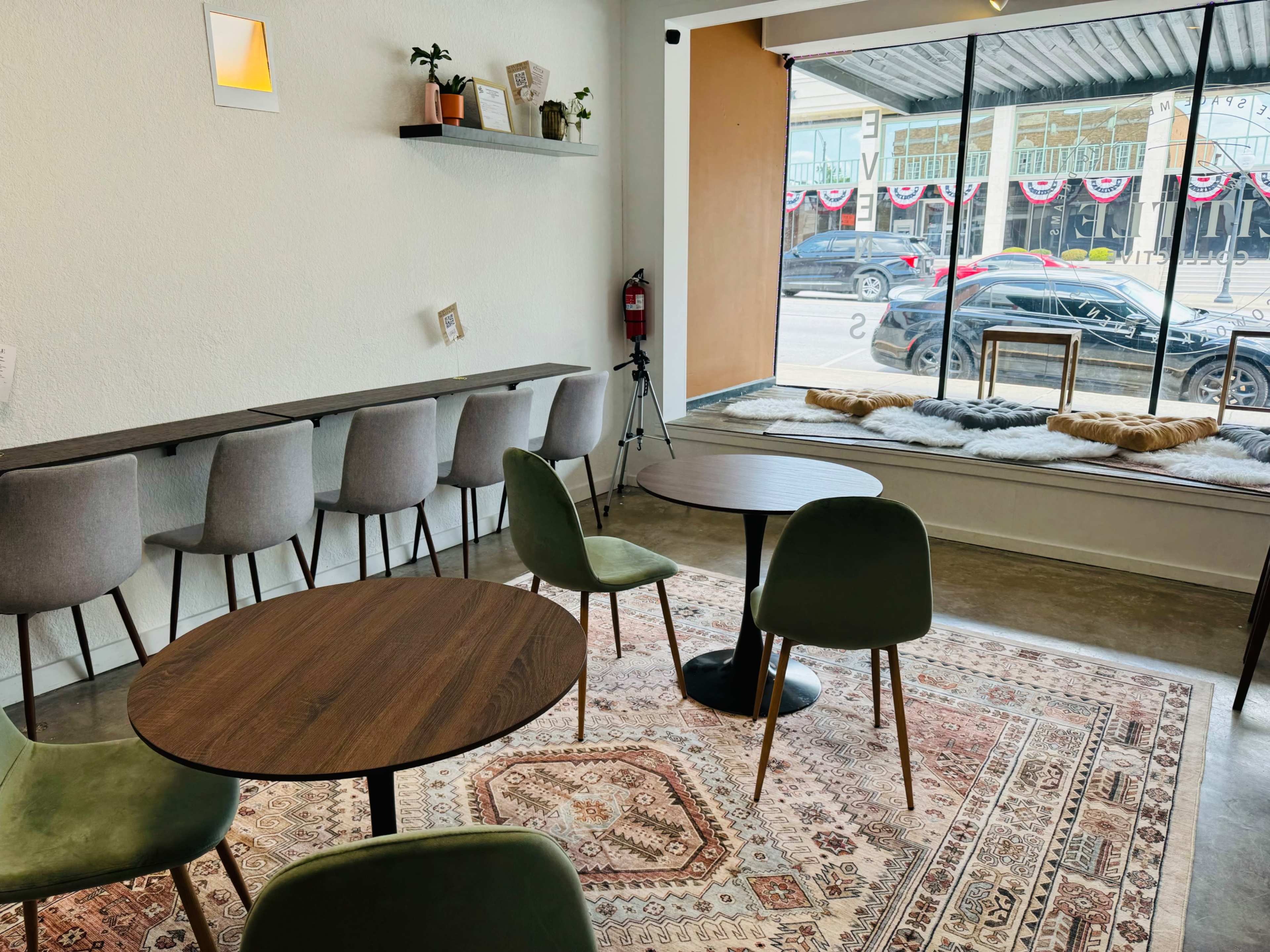 A cozy café interior with several tables and chairs arranged on a patterned rug, overlooking a street through a large window.