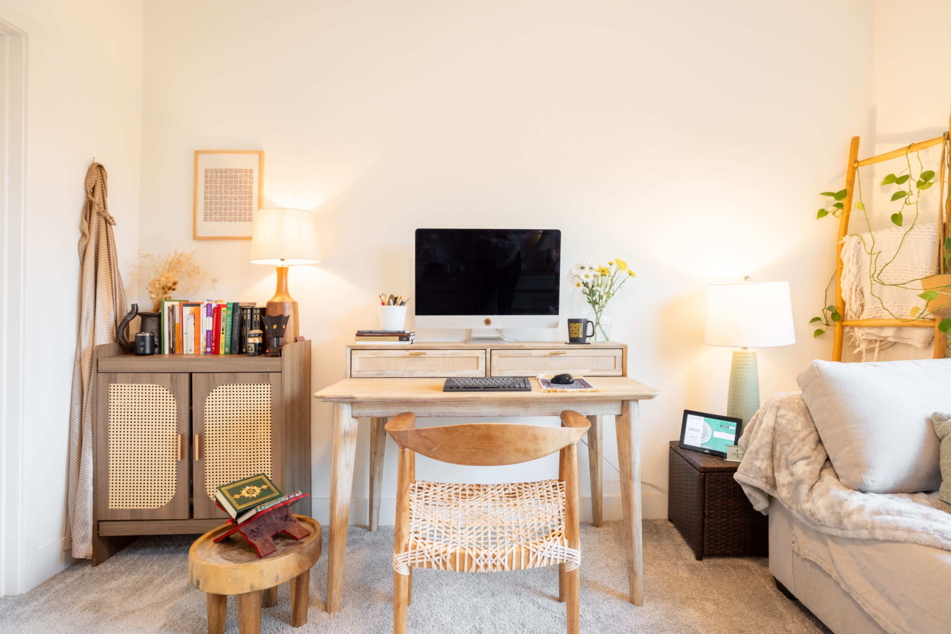 The image shows a brightly lit home office with a wooden desk, an iMac computer, and a cozy seating area featuring a light-colored sofa.