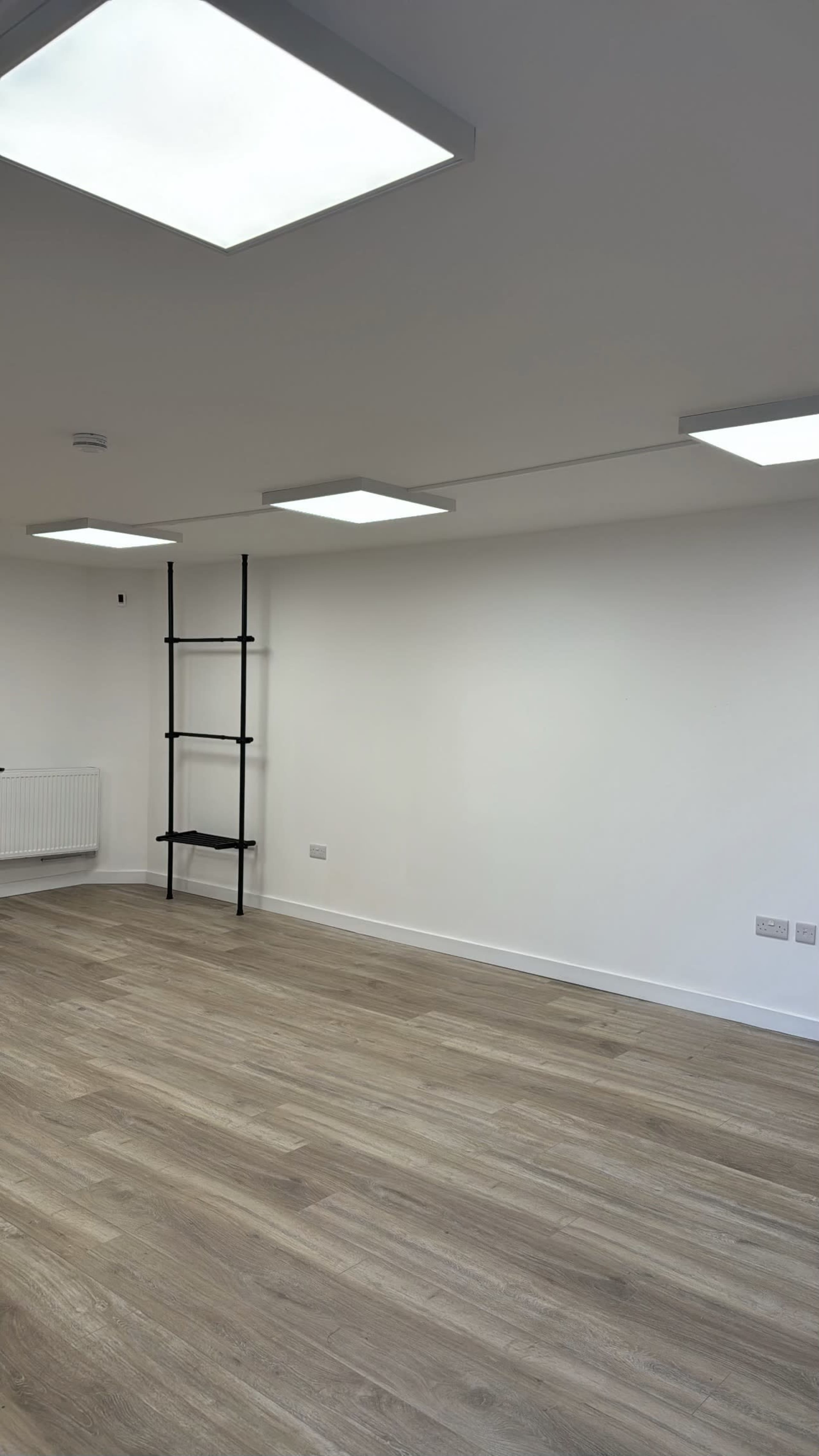 The image shows a minimalist room with wooden flooring, a white wall, a ladder shelf, and bright ceiling lights.