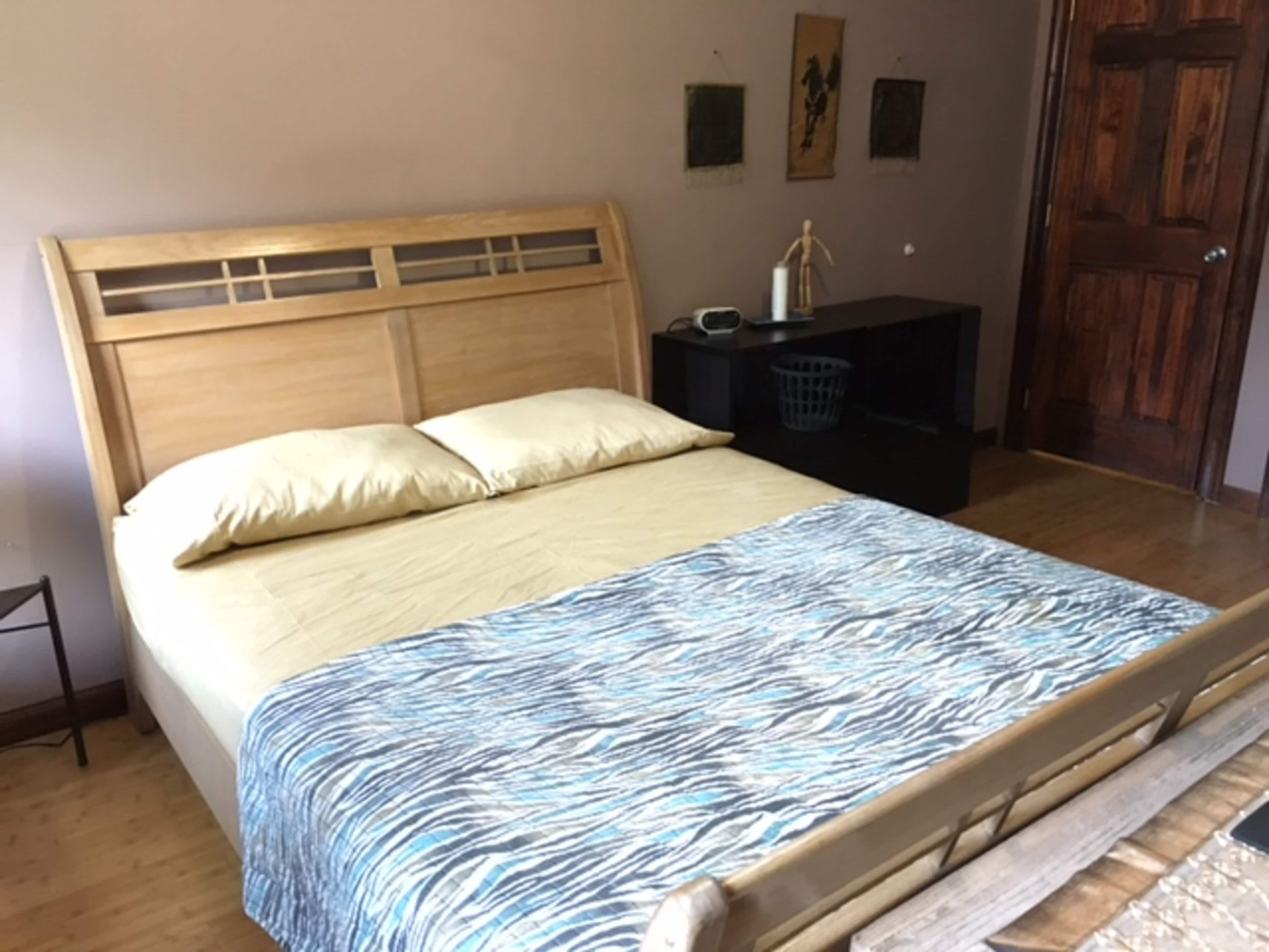 A wooden bed with beige pillows and a patterned blanket is positioned in a room with a dark wooden door and wall art.