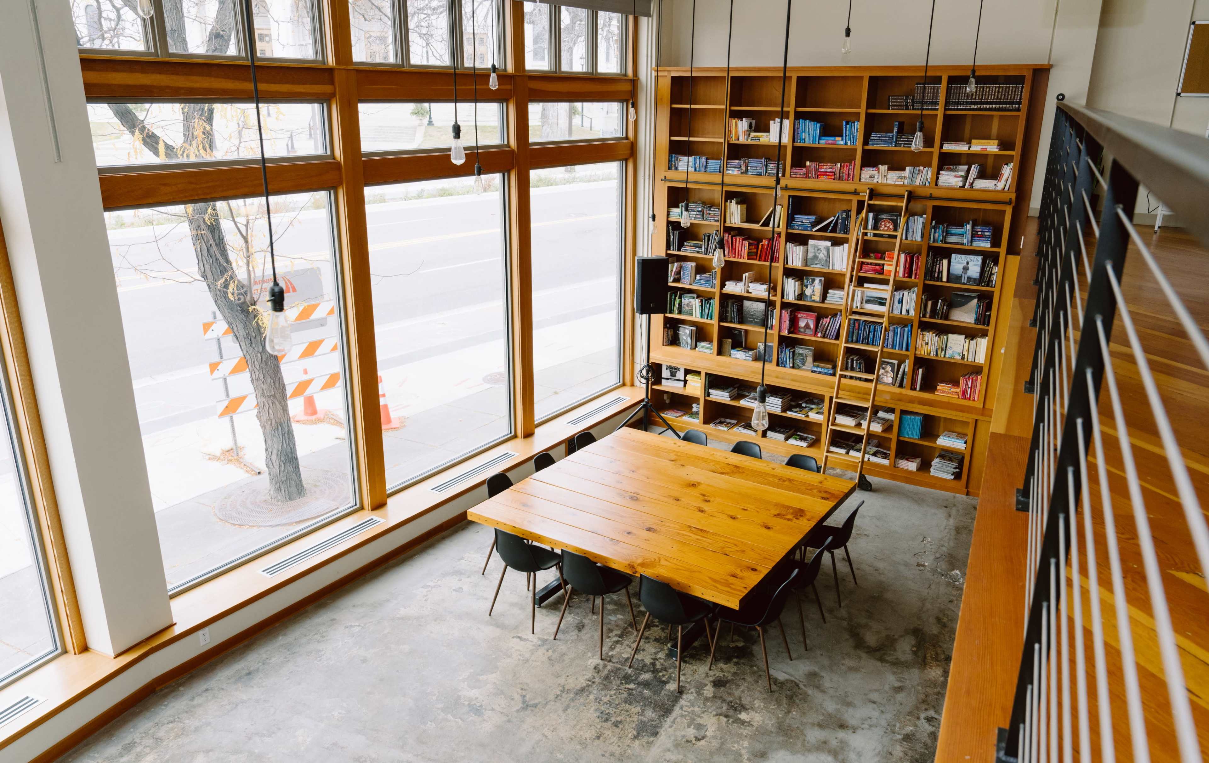 The image shows a large wooden table surrounded by chairs in a well-lit room with bookshelves and large windows facing a street.