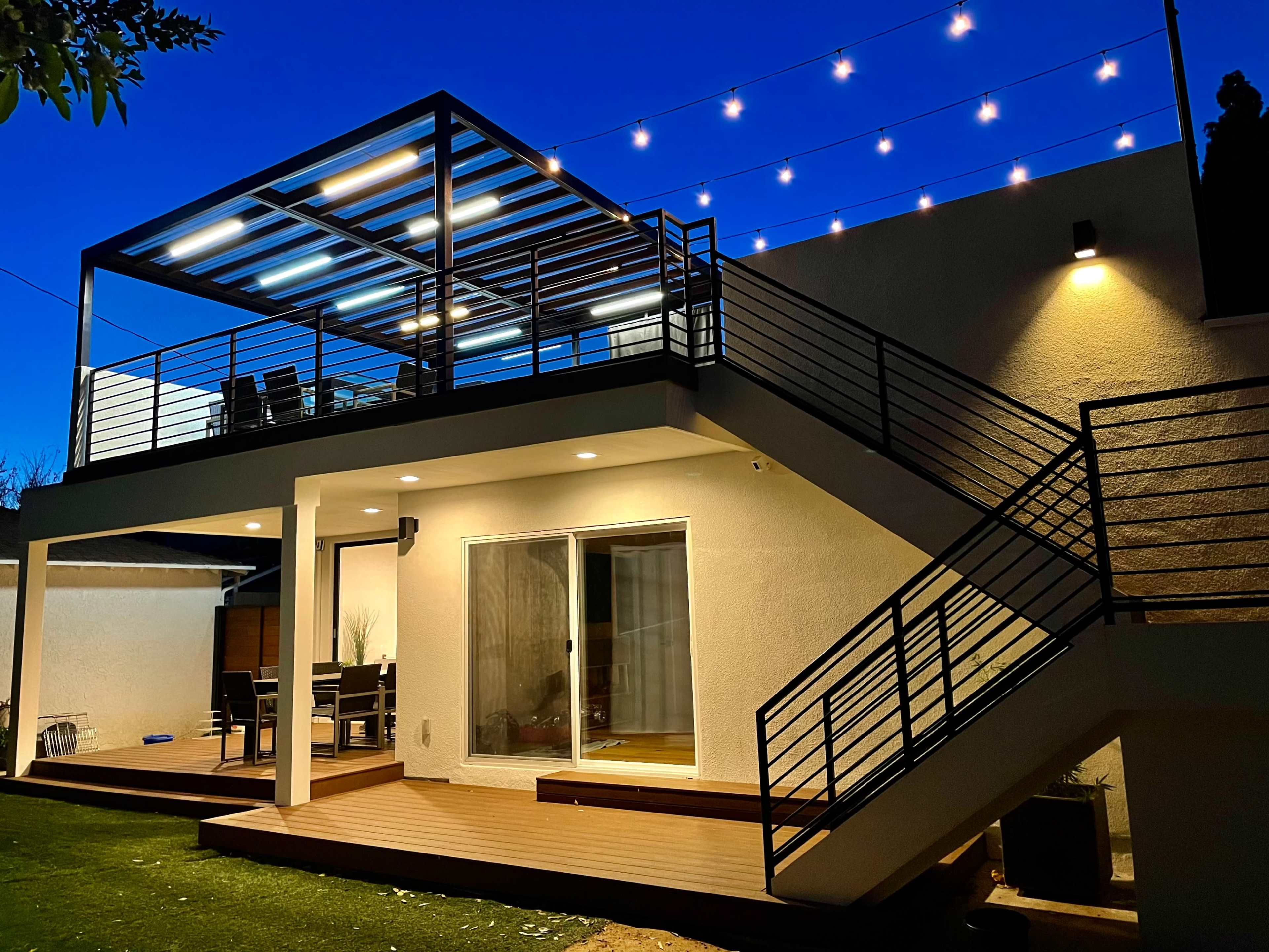 A modern two-story house features a spacious deck illuminated by string lights and a winding staircase leading to the upper level.