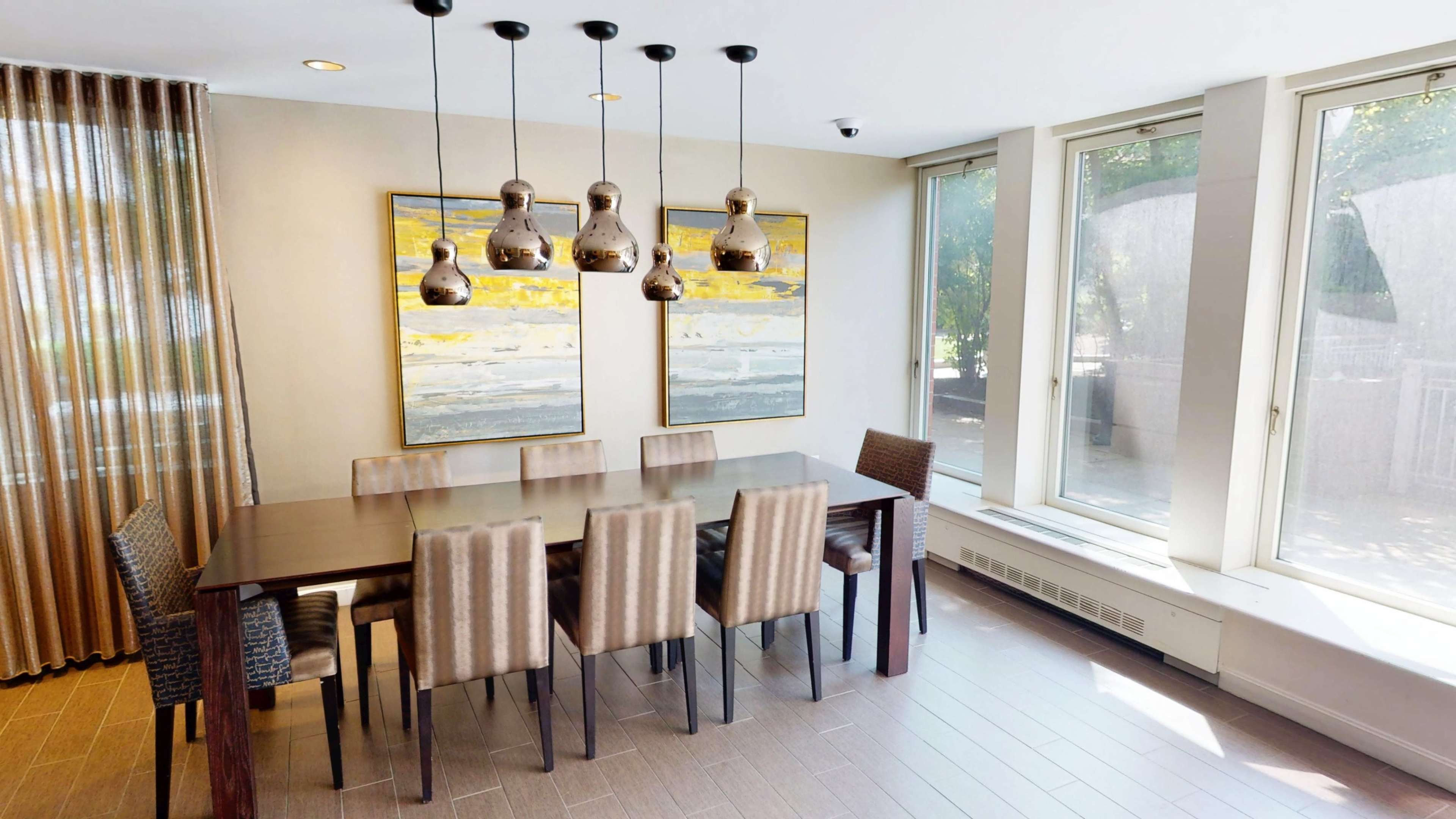 The image shows a modern dining area with a long wooden table surrounded by eight chairs, two abstract paintings on the wall, and pendant lights hanging above.