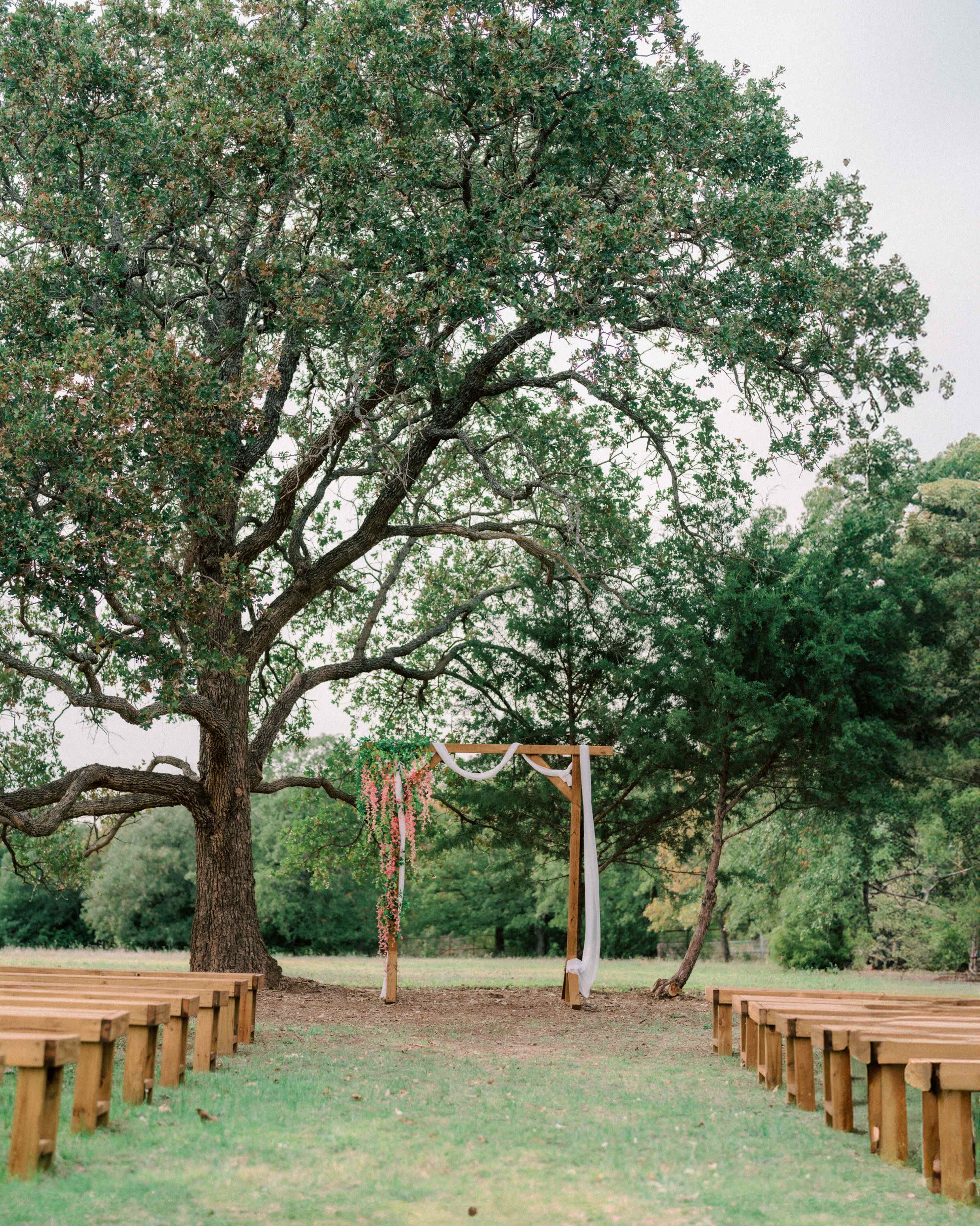An outdoor wedding ceremony setting features wooden benches arranged in rows facing a floral-adorned arch under a large tree.
