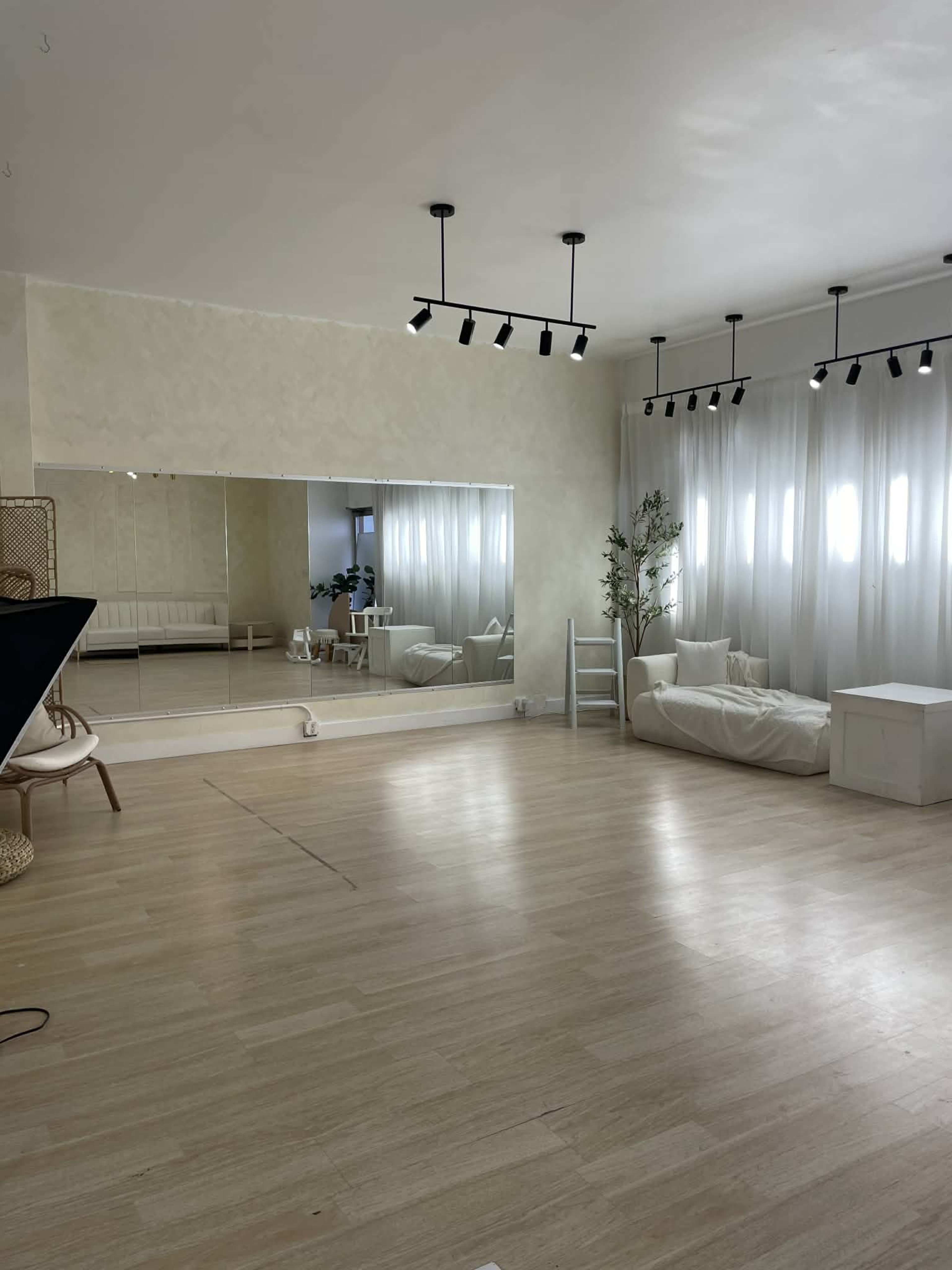 The image shows a bright, spacious studio with mirrored walls, large windows, and minimalist furniture.