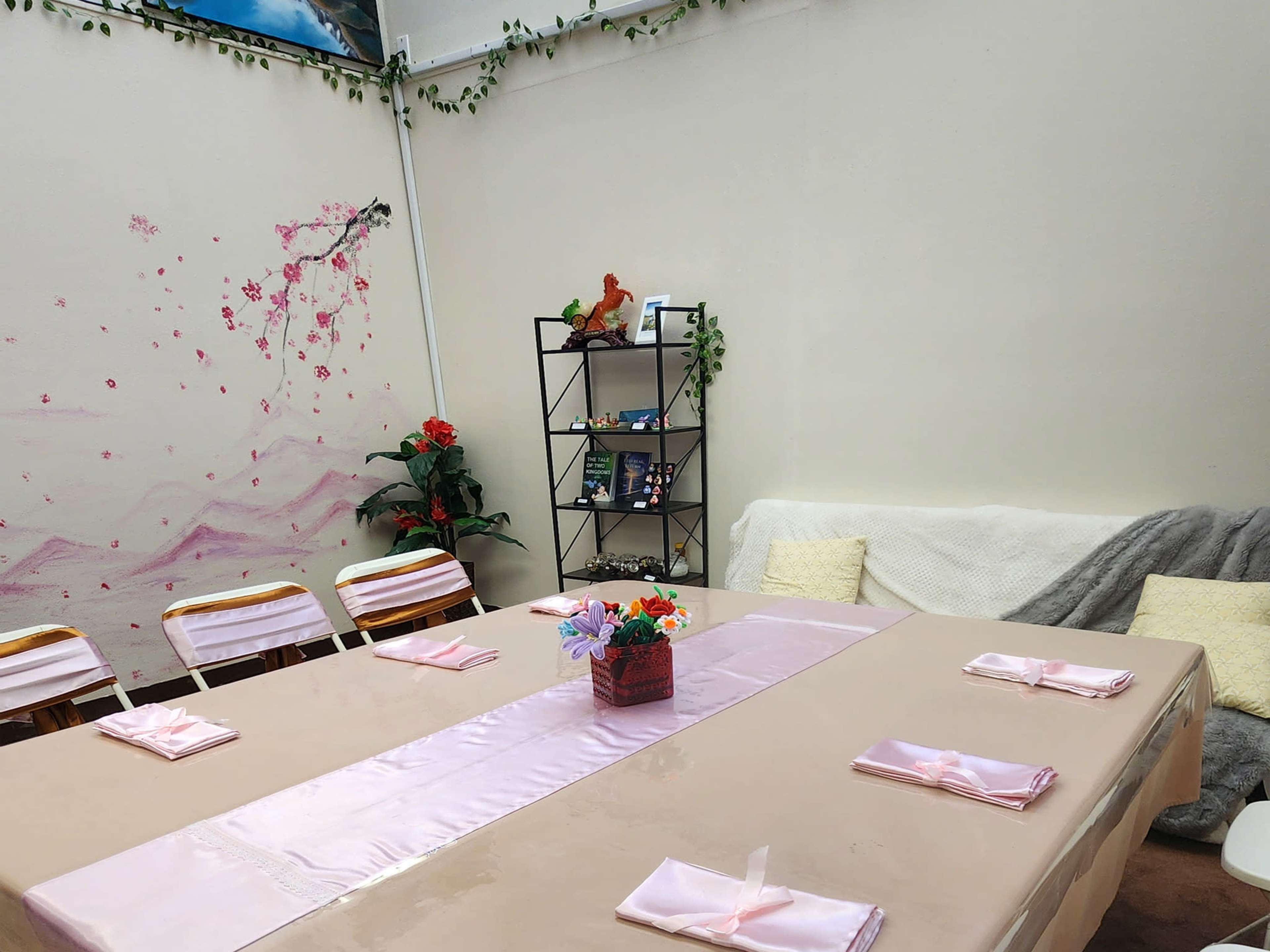 The image shows a decorated room with a long table set with pink cloth and napkins, surrounded by chairs, and features a shelf with a few decorative items and plants.