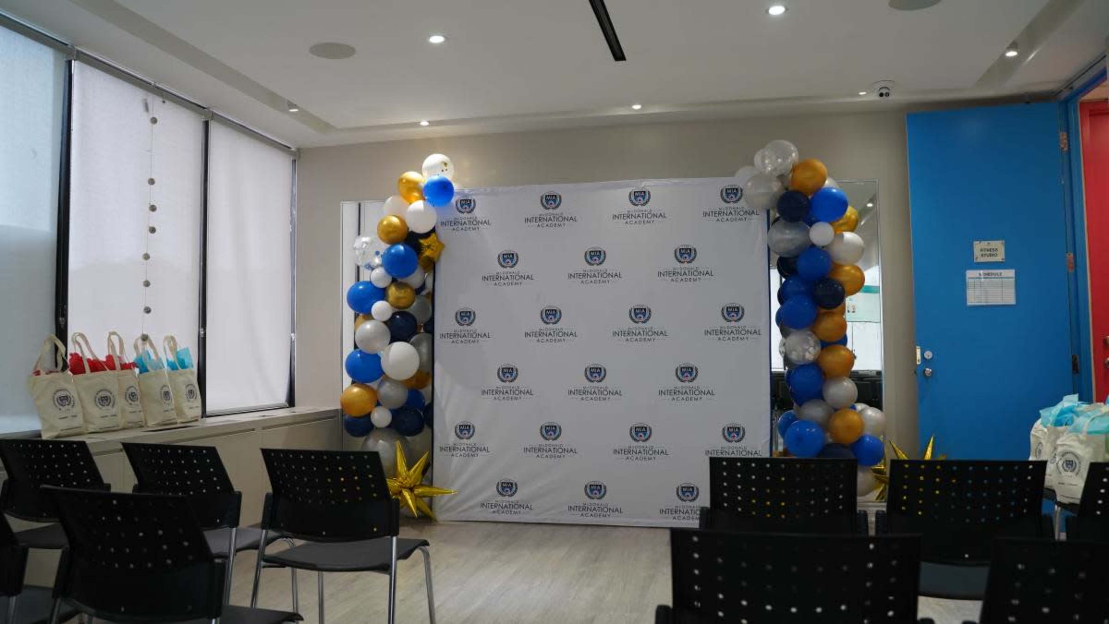 The image shows a decorated event space with a backdrop featuring a logo, flanked by clusters of balloons in blue, white, and gold, and chairs arranged in rows facing the backdrop.