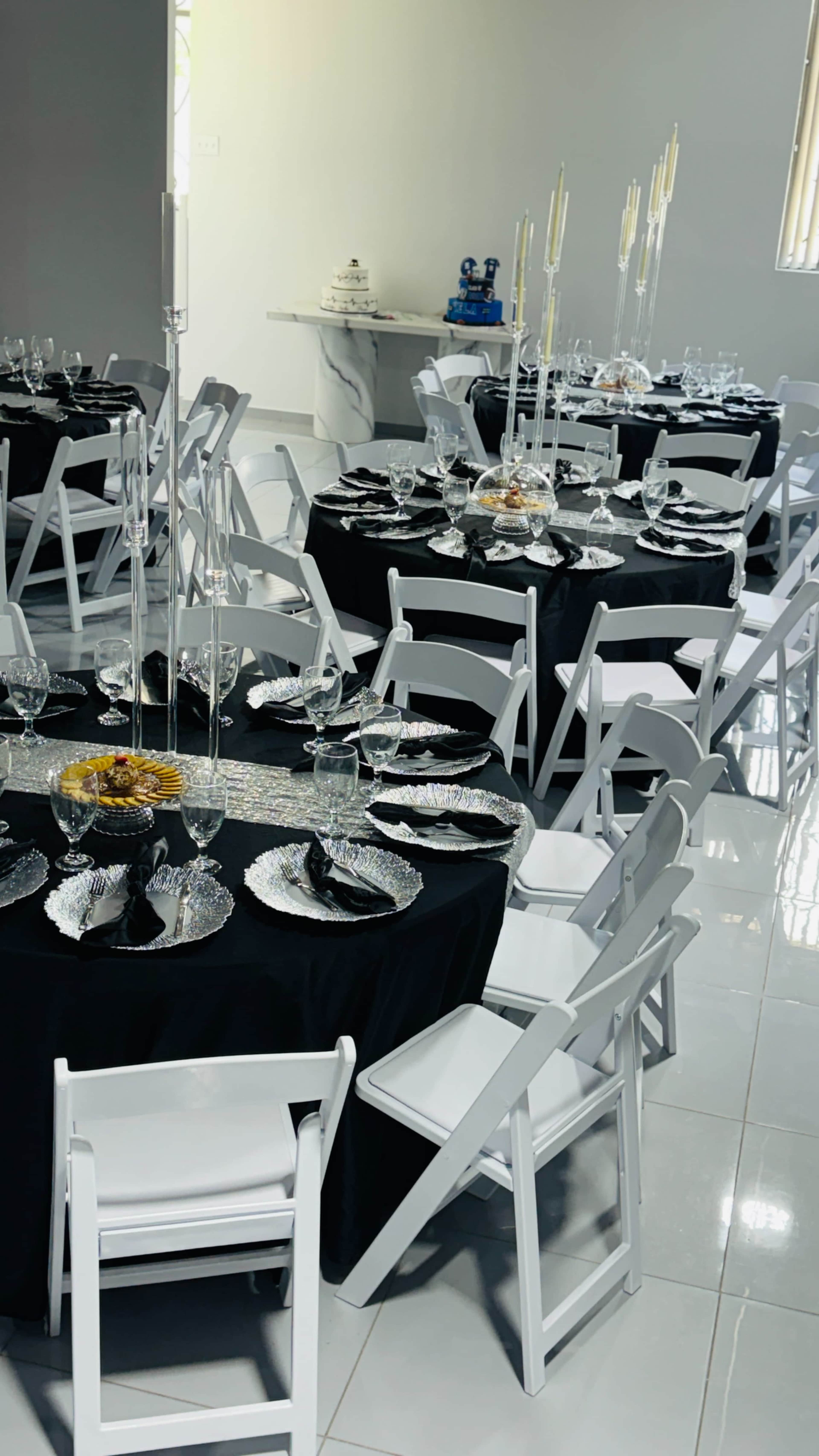 The image shows a banquet hall setup with round tables covered in black tablecloths, each adorned with elegant dinnerware and centerpieces, surrounded by white folding chairs.