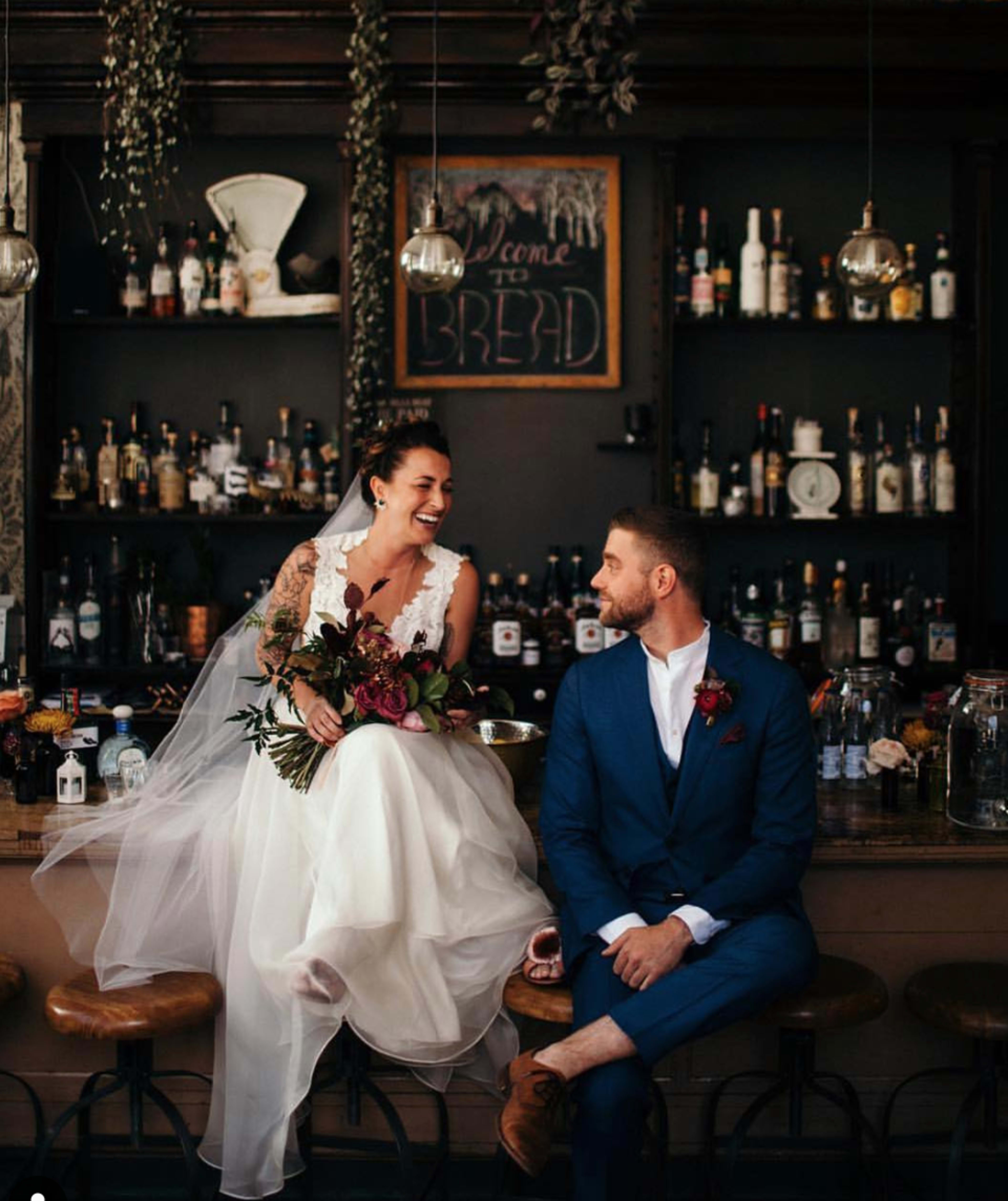 A bride and groom sit at a bar, with the bride perched on the counter and holding a bouquet, while the groom looks at her.