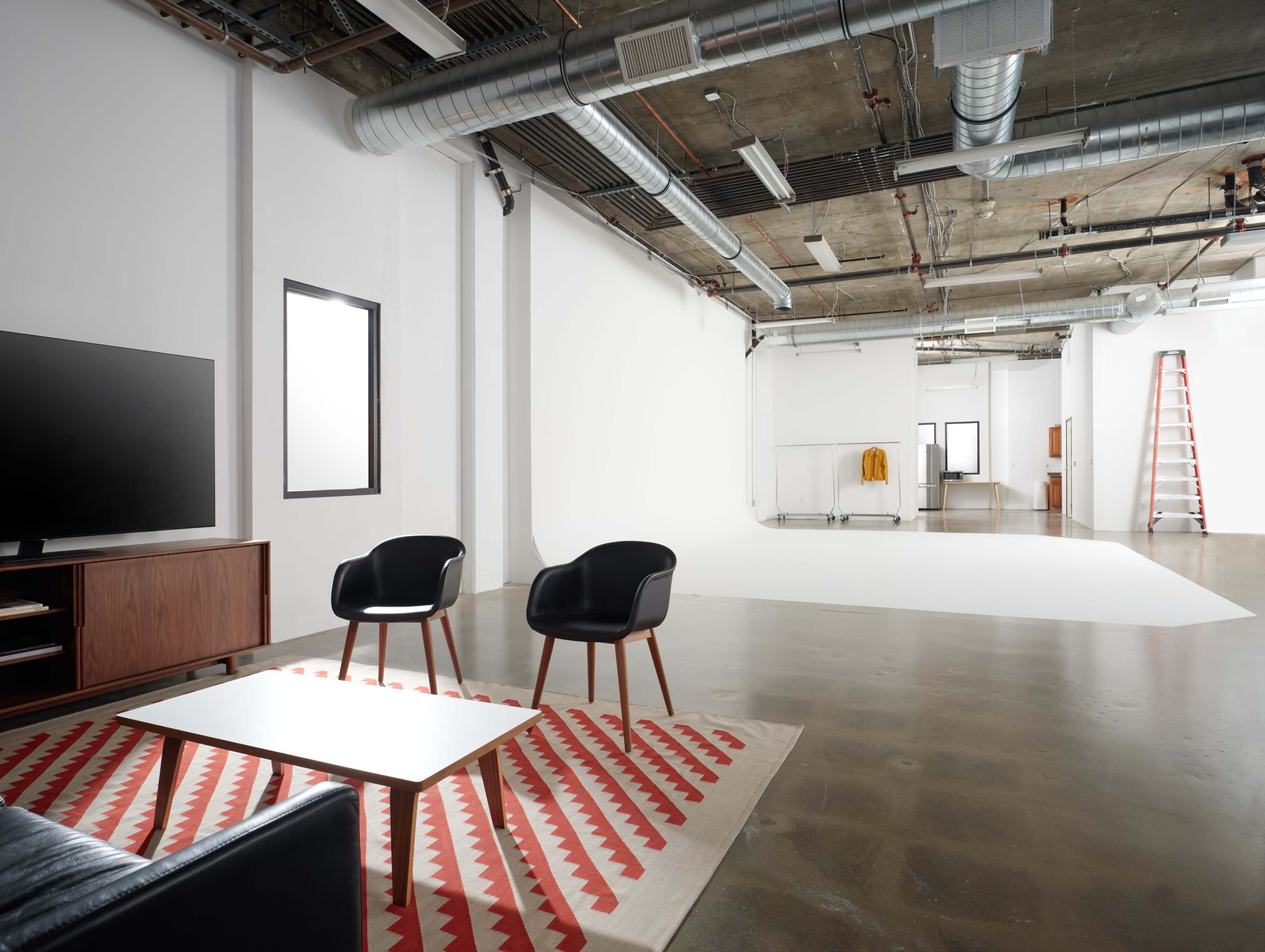A modern studio space featuring a black couch, two chairs, a coffee table on a patterned rug, and a ladder in the background.
