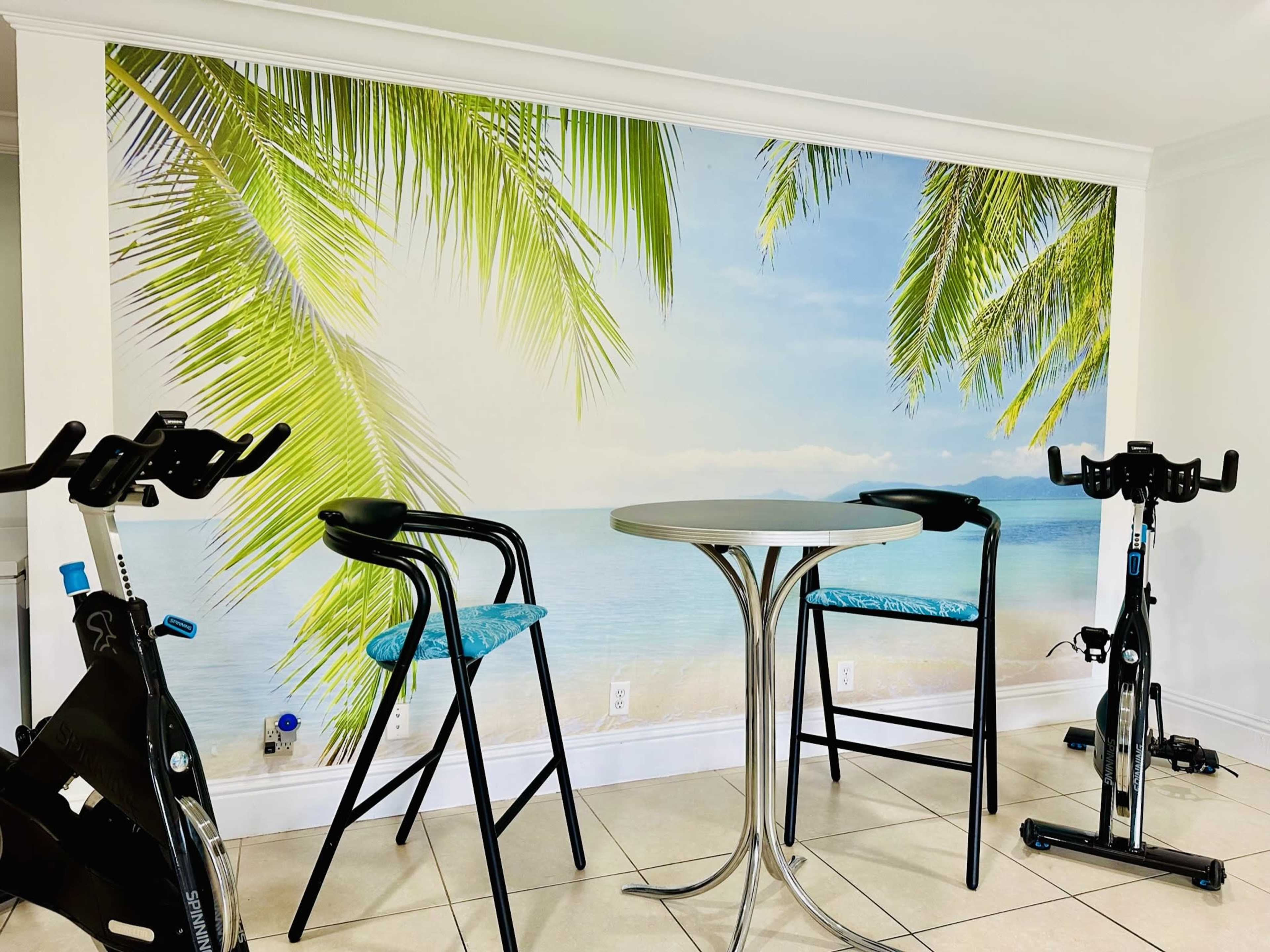 A bright room with a beach-themed mural featuring palm leaves, a round table with two bar stools, and stationary exercise bikes positioned nearby.