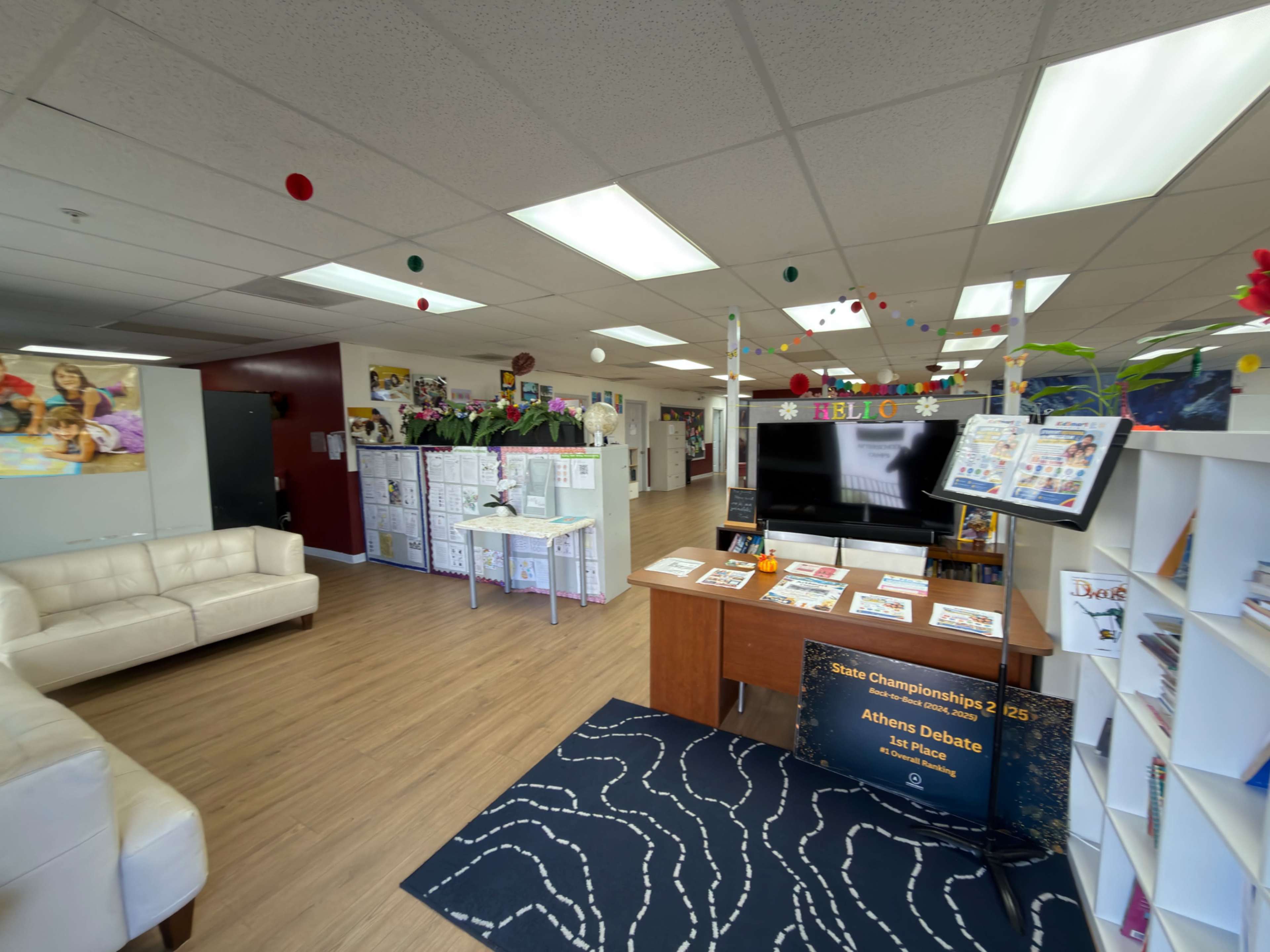 The image shows a brightly lit community center interior with a reception area featuring a desk, a television, and seating arrangements, along with shelves displaying magazines and informational materials.