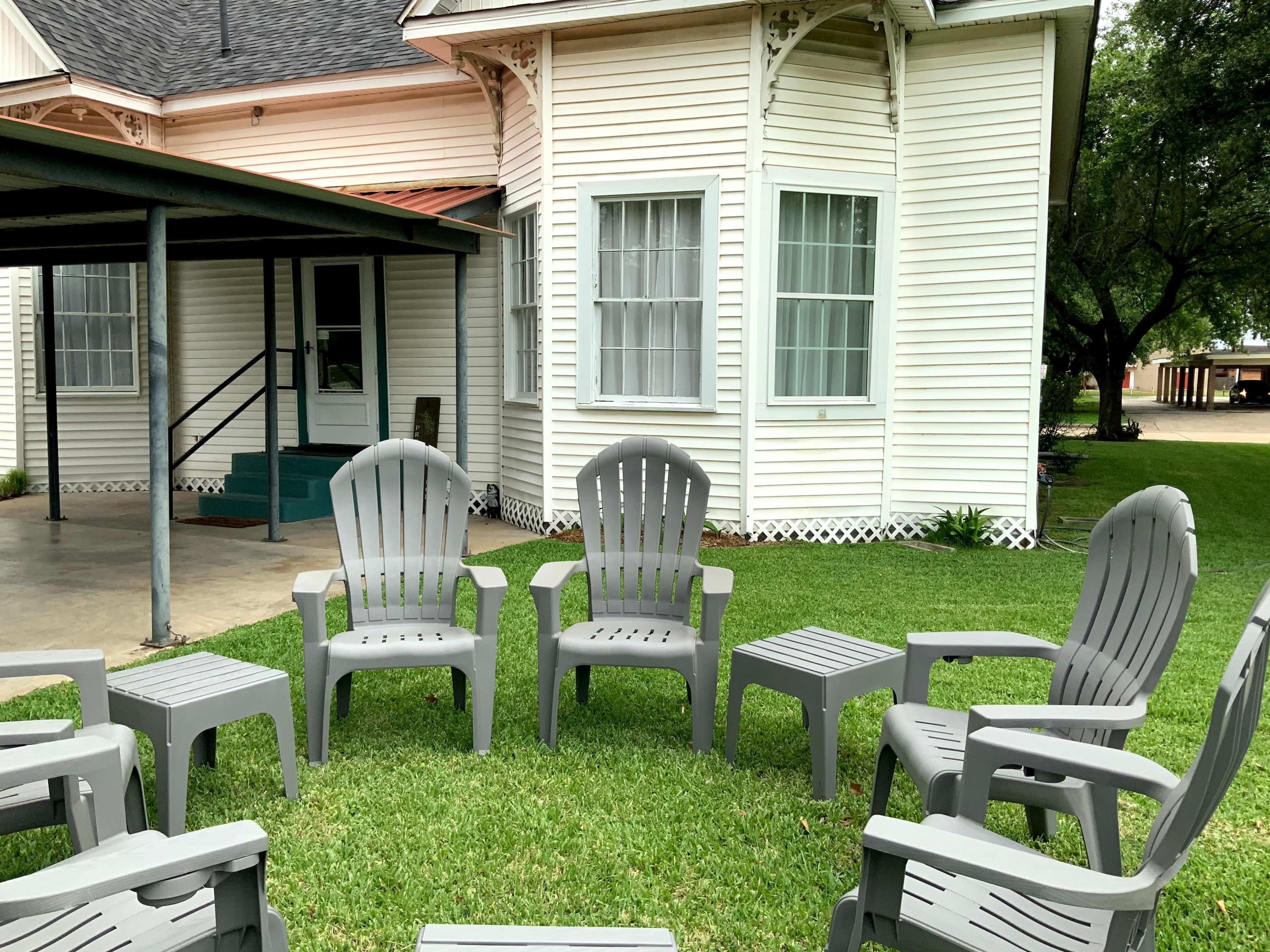 A set of gray plastic chairs is arranged in a circle on a grassy area in front of a white house.