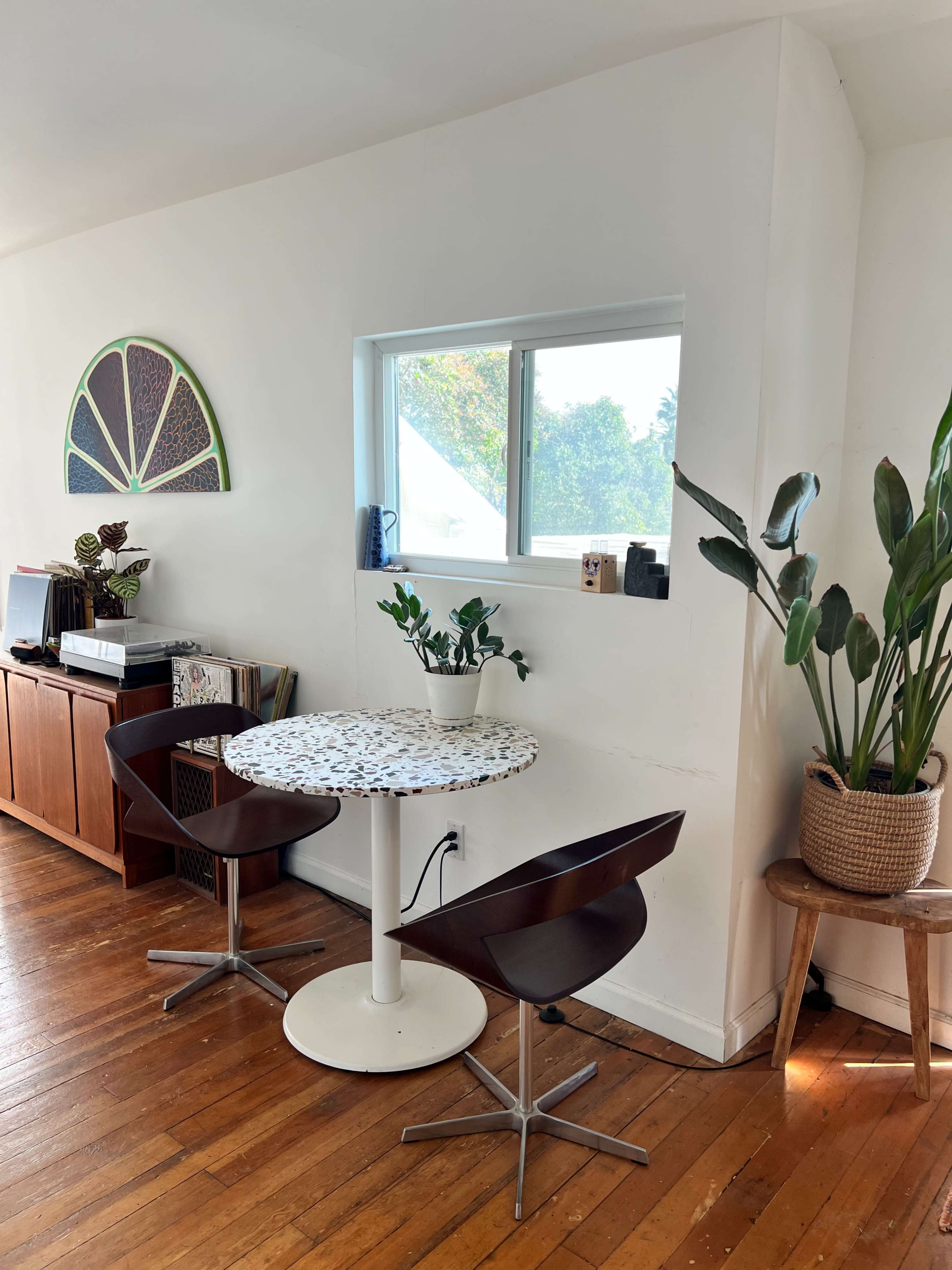 A small dining area features a round table with a speckled surface and two chairs, next to a window and surrounded by indoor plants and wooden furniture.