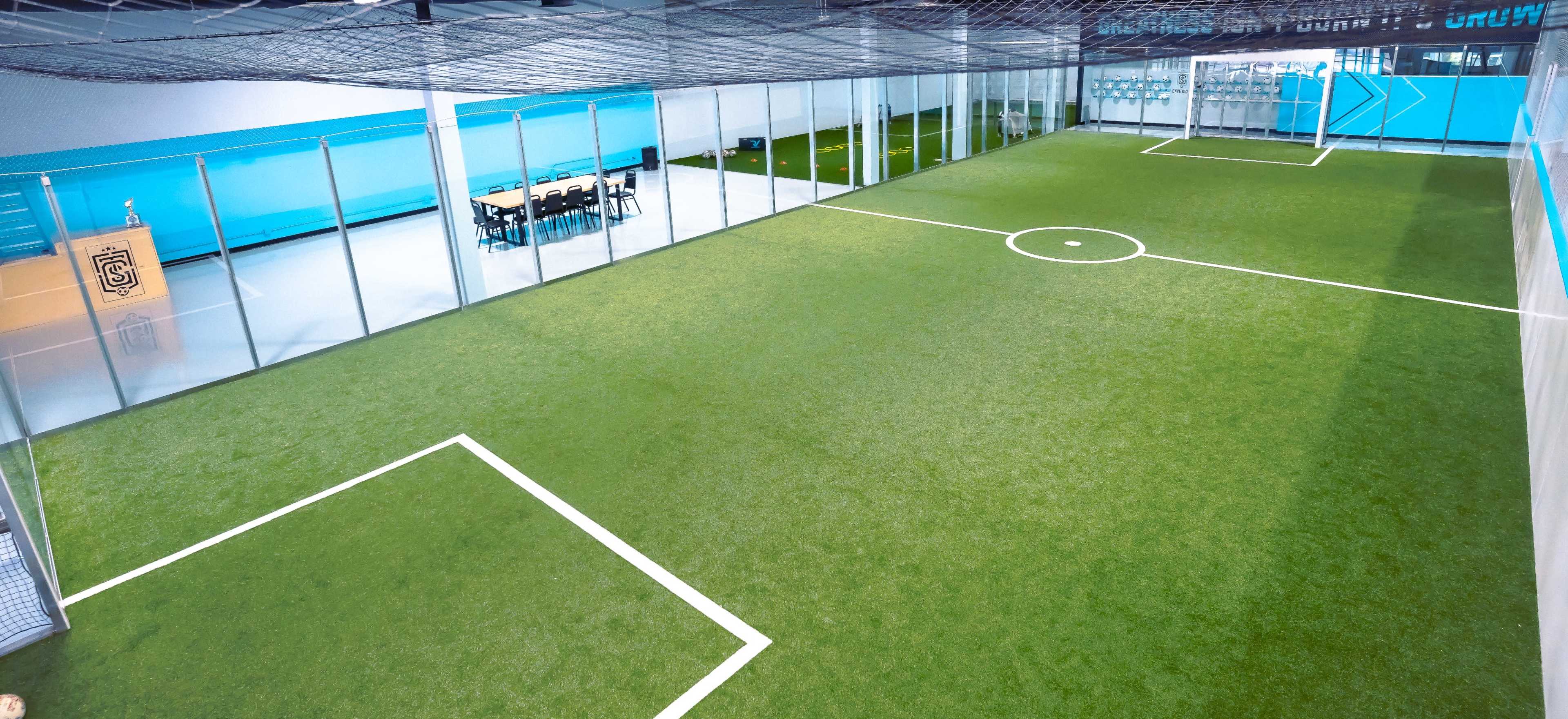 The image shows a small indoor soccer field surrounded by glass walls, featuring a central goal and seating area at one end.