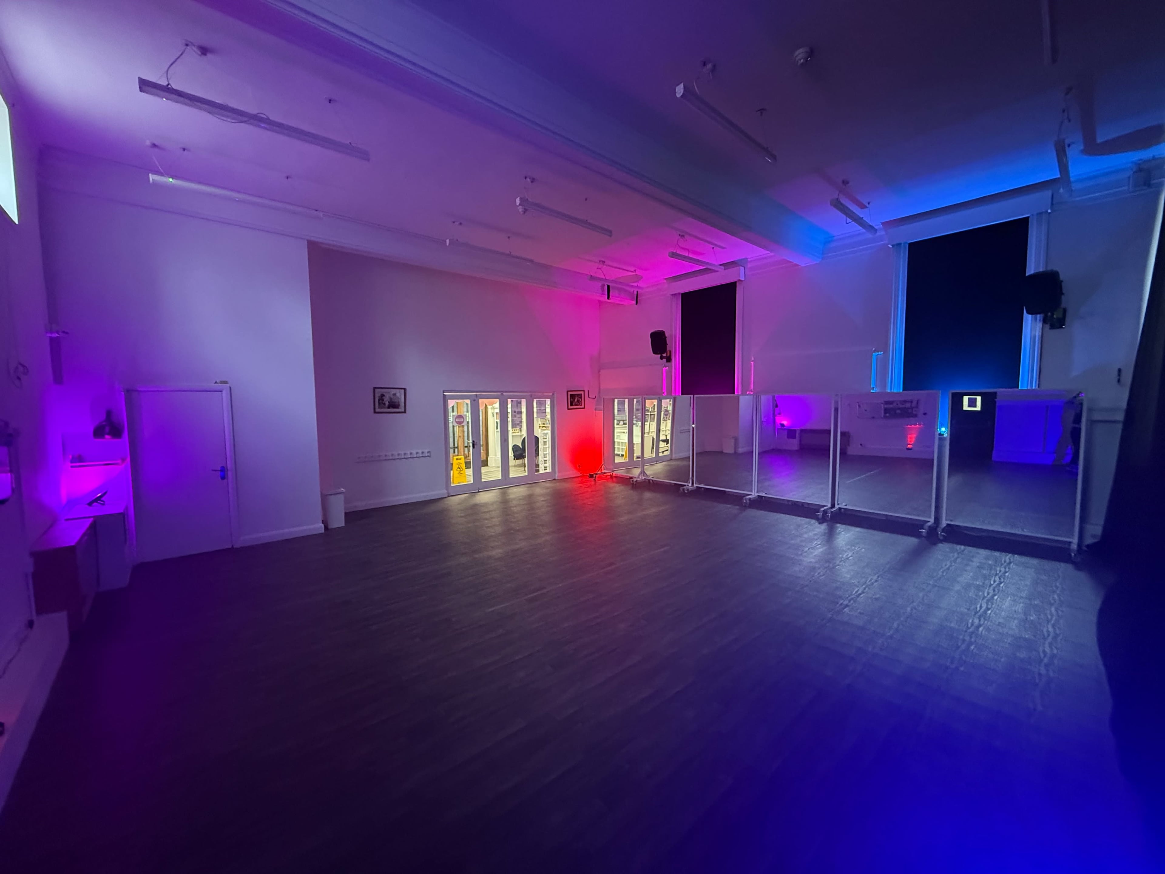 The image shows a spacious room with modern lighting in shades of purple and red, featuring glass partitions and large windows.