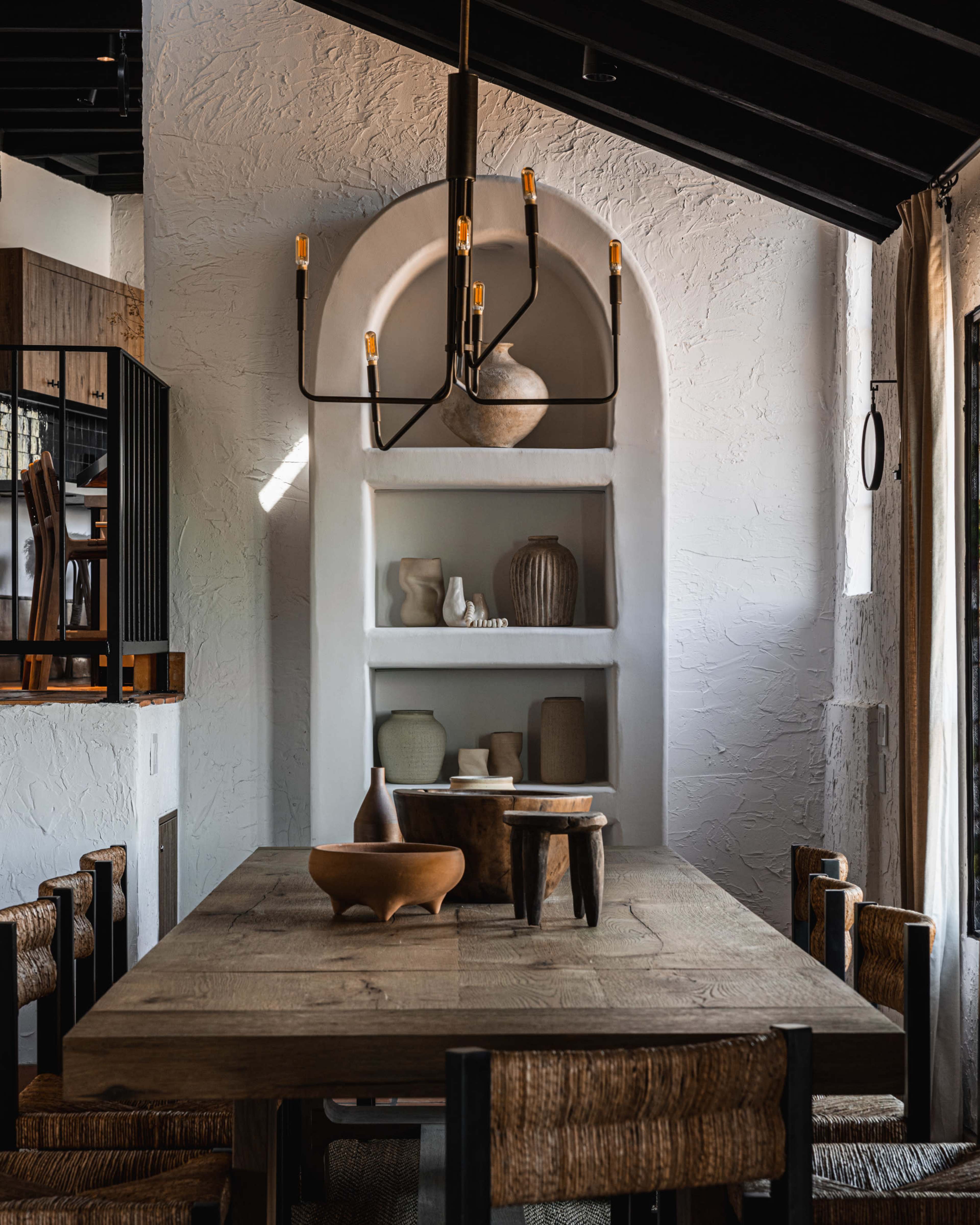 The image depicts a rustic dining area featuring a wooden table surrounded by woven chairs, with a prominent light fixture hanging above and a display shelf with ceramic and stone objects in the background.