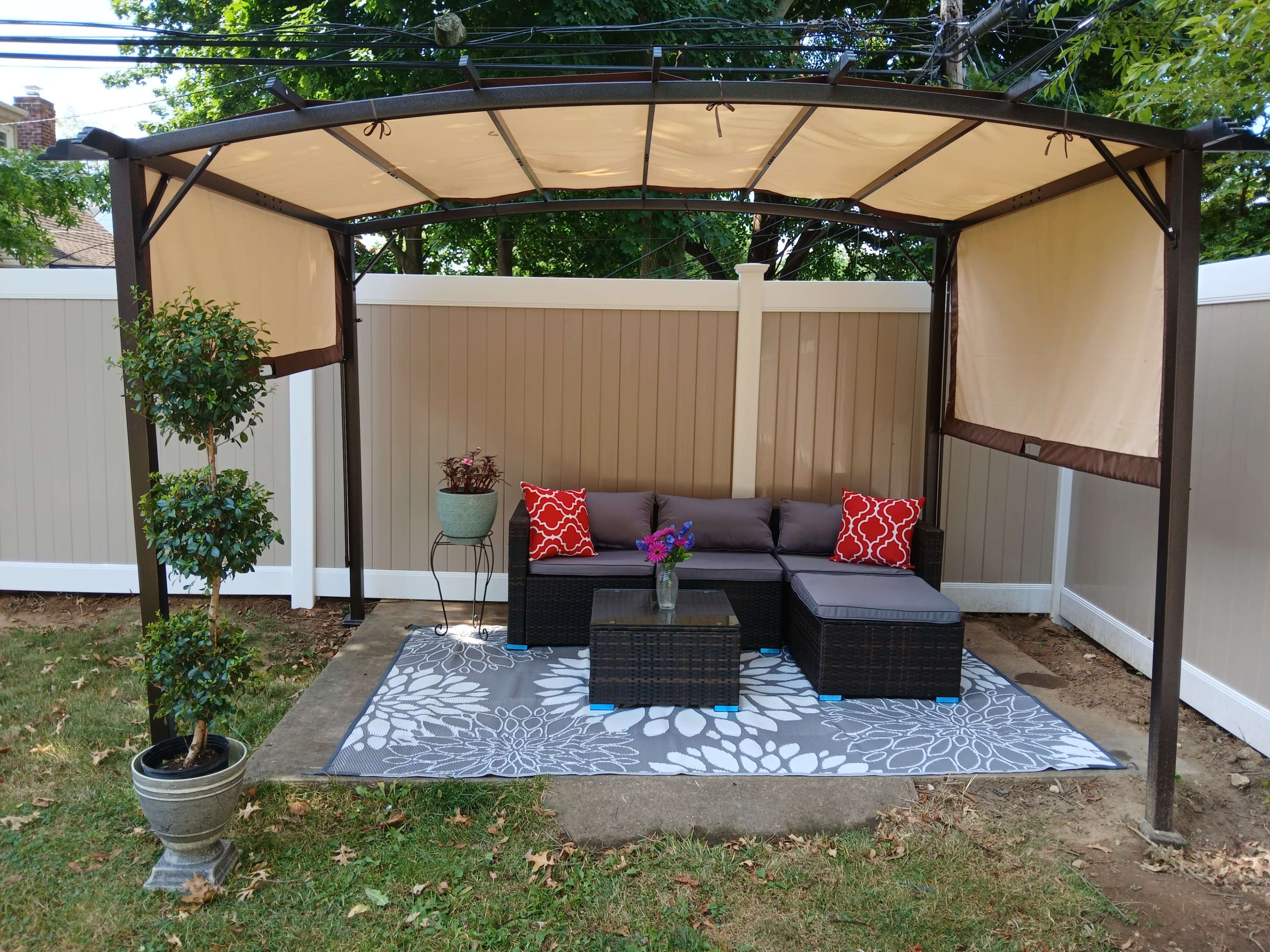 A metal gazebo with a tan canopy covers a seating area consisting of a sofa, a coffee table, and decorative cushions, set on a patterned rug in a backyard.