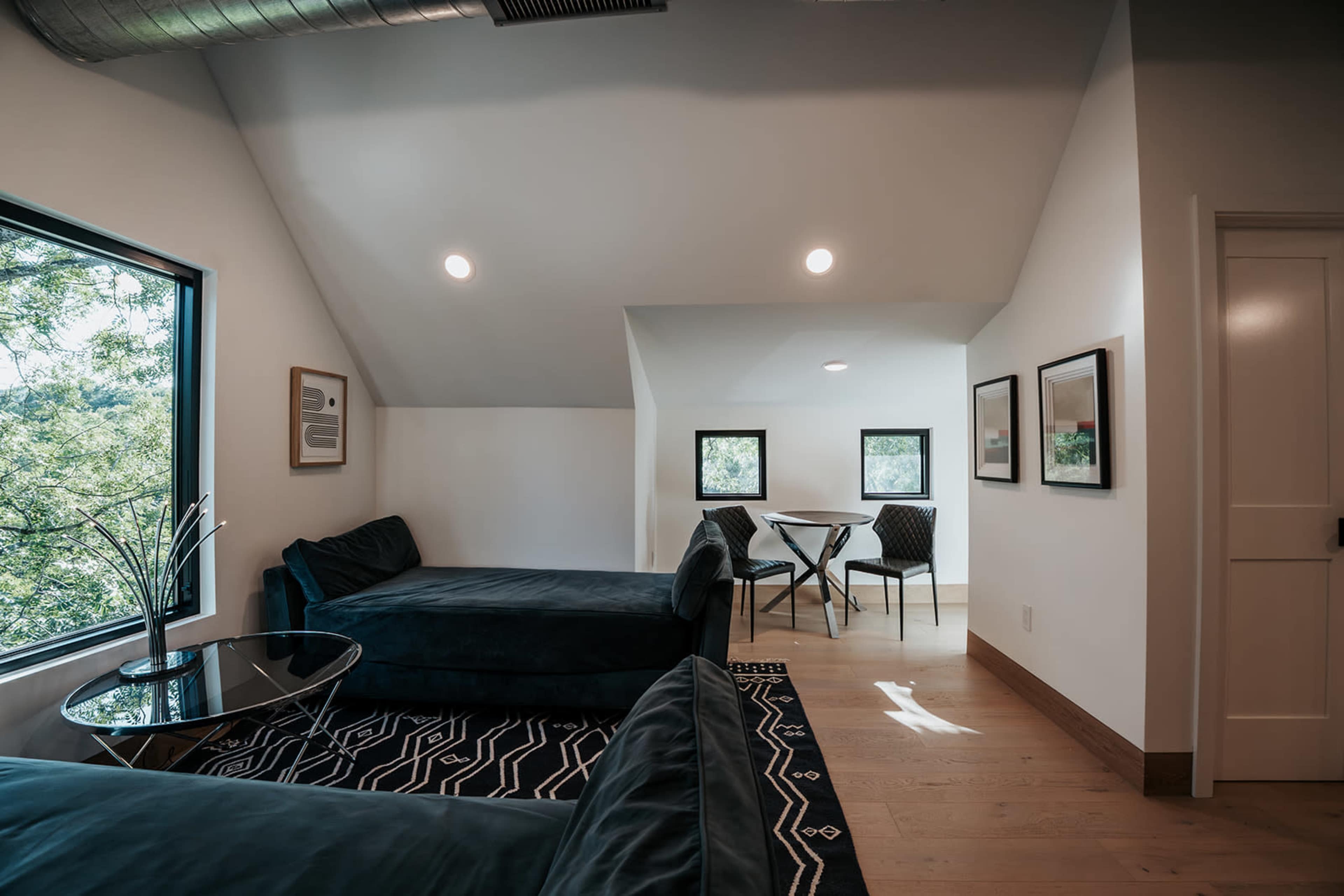 A well-lit room features two dark couches, a glass side table, and a dining set with four chairs, flanked by three small windows.