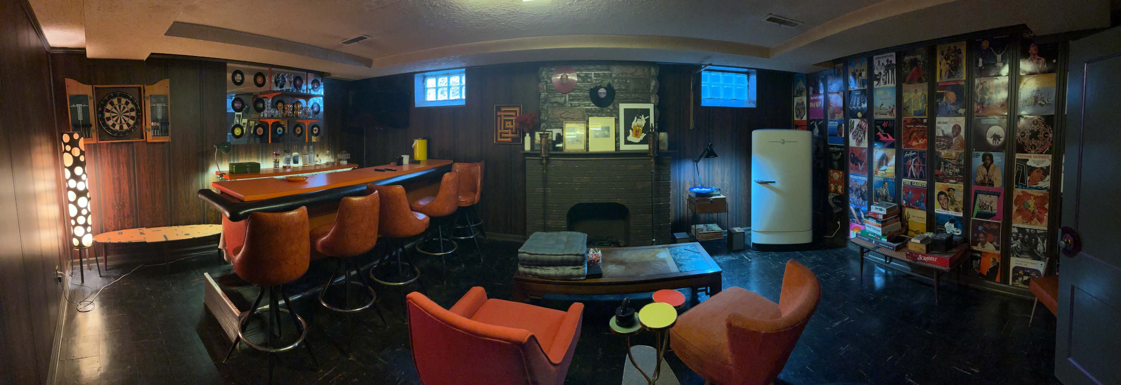 The image shows a basement lounge with a bar area, dartboard, vinyl record collection on the walls, and vintage furniture, including two orange chairs and a retro refrigerator.
