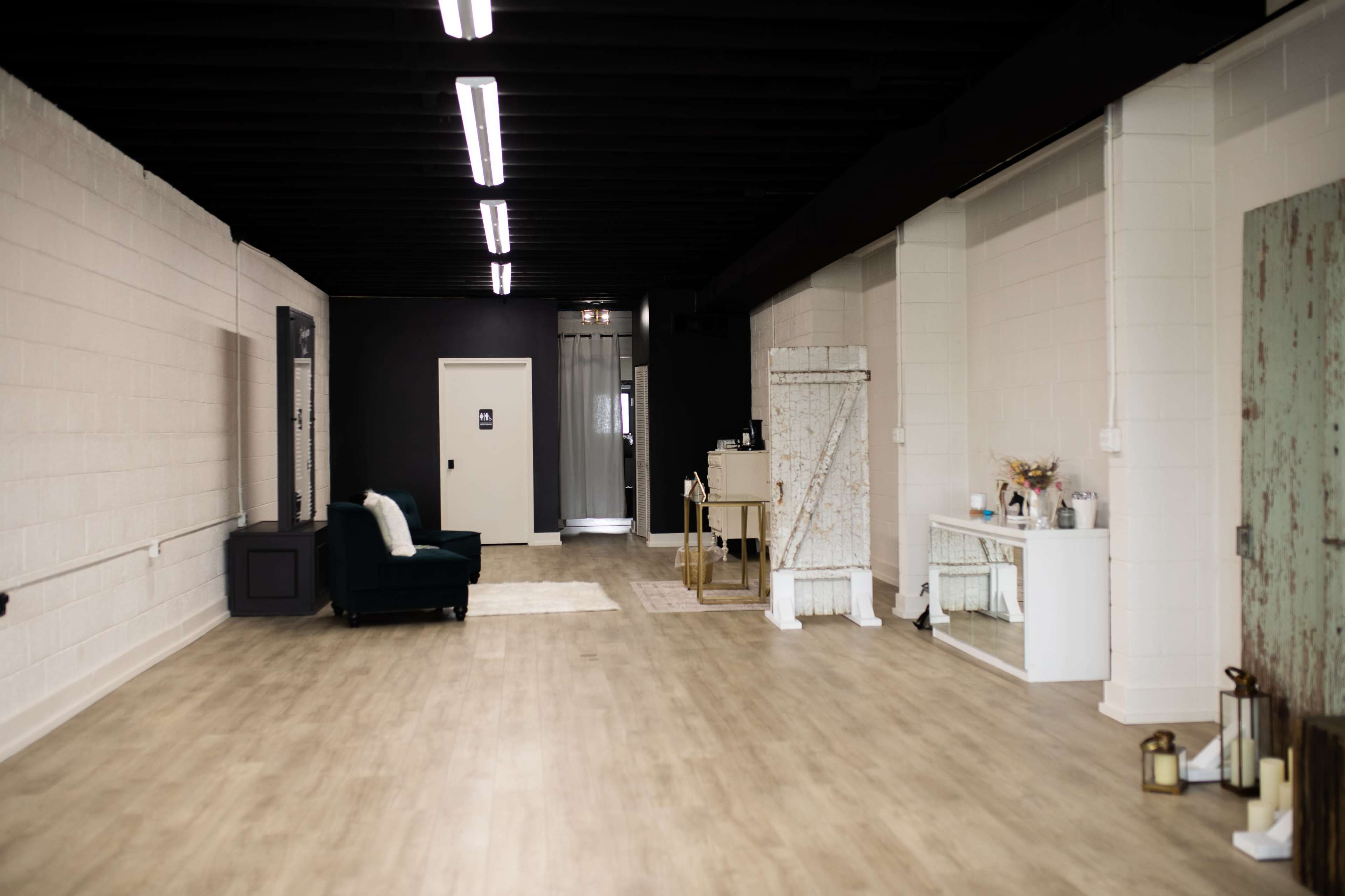 The image shows a spacious interior with light-colored walls, a wooden floor, and various furniture pieces along the sides, including a green chair, a small table, and a large mirror.