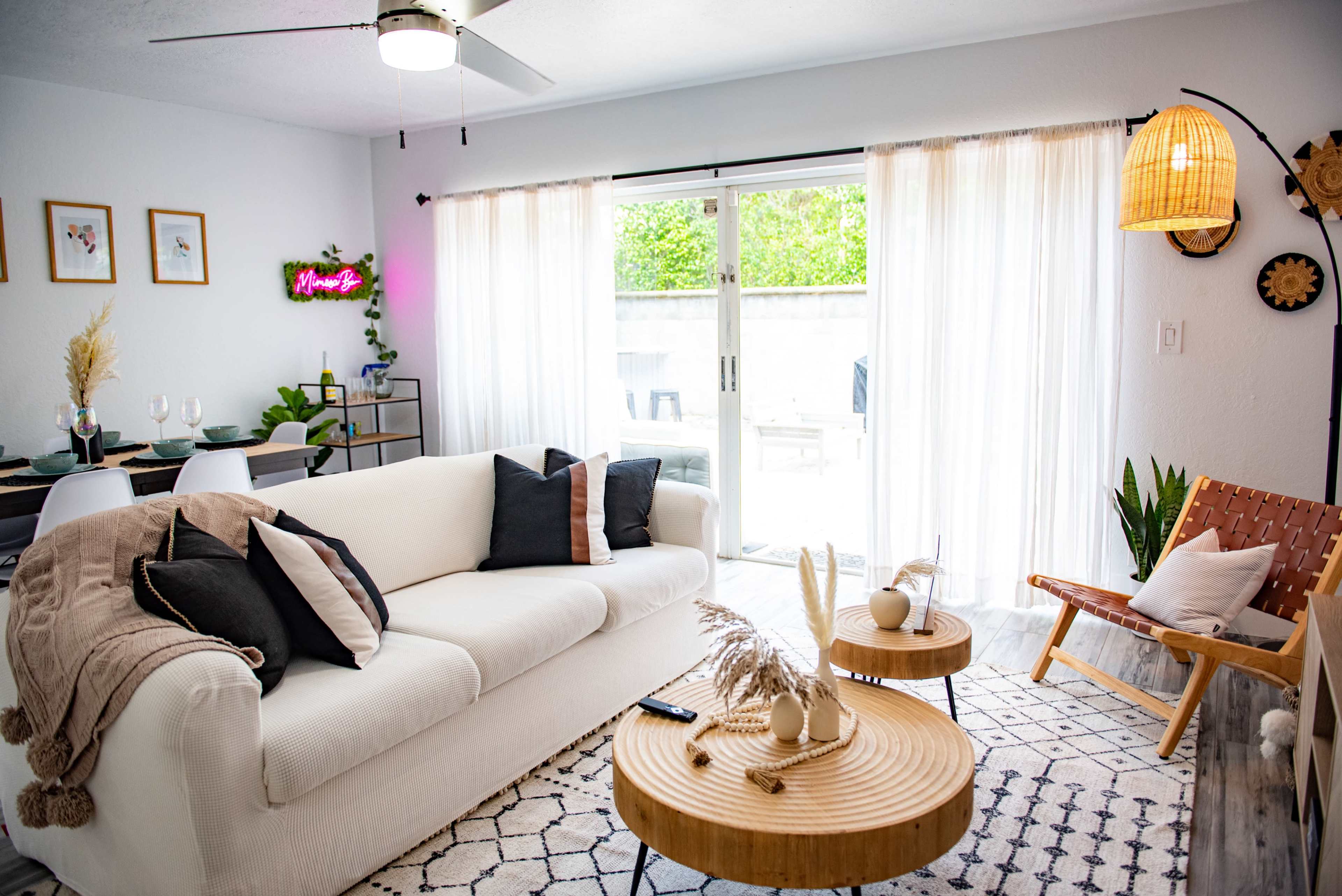 A bright living space featuring a white sofa with black and beige pillows, wooden coffee tables, and large glass doors leading to a patio, all decorated with plants and light fixtures.