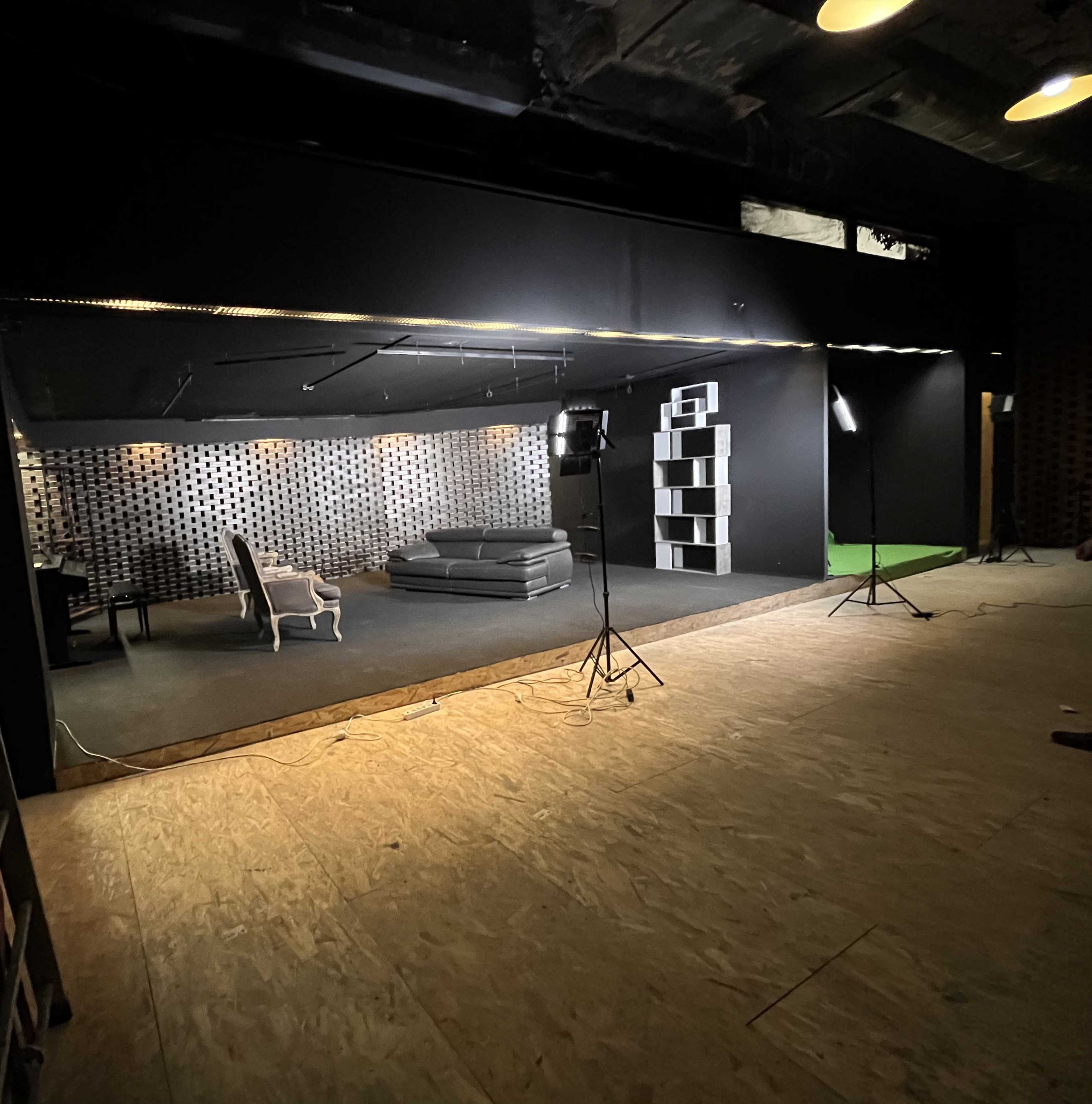 The image shows a dimly lit studio with a black backdrop, featuring a gray sofa, a white chair, and a bookshelf against the wall, all set on a wooden floor.