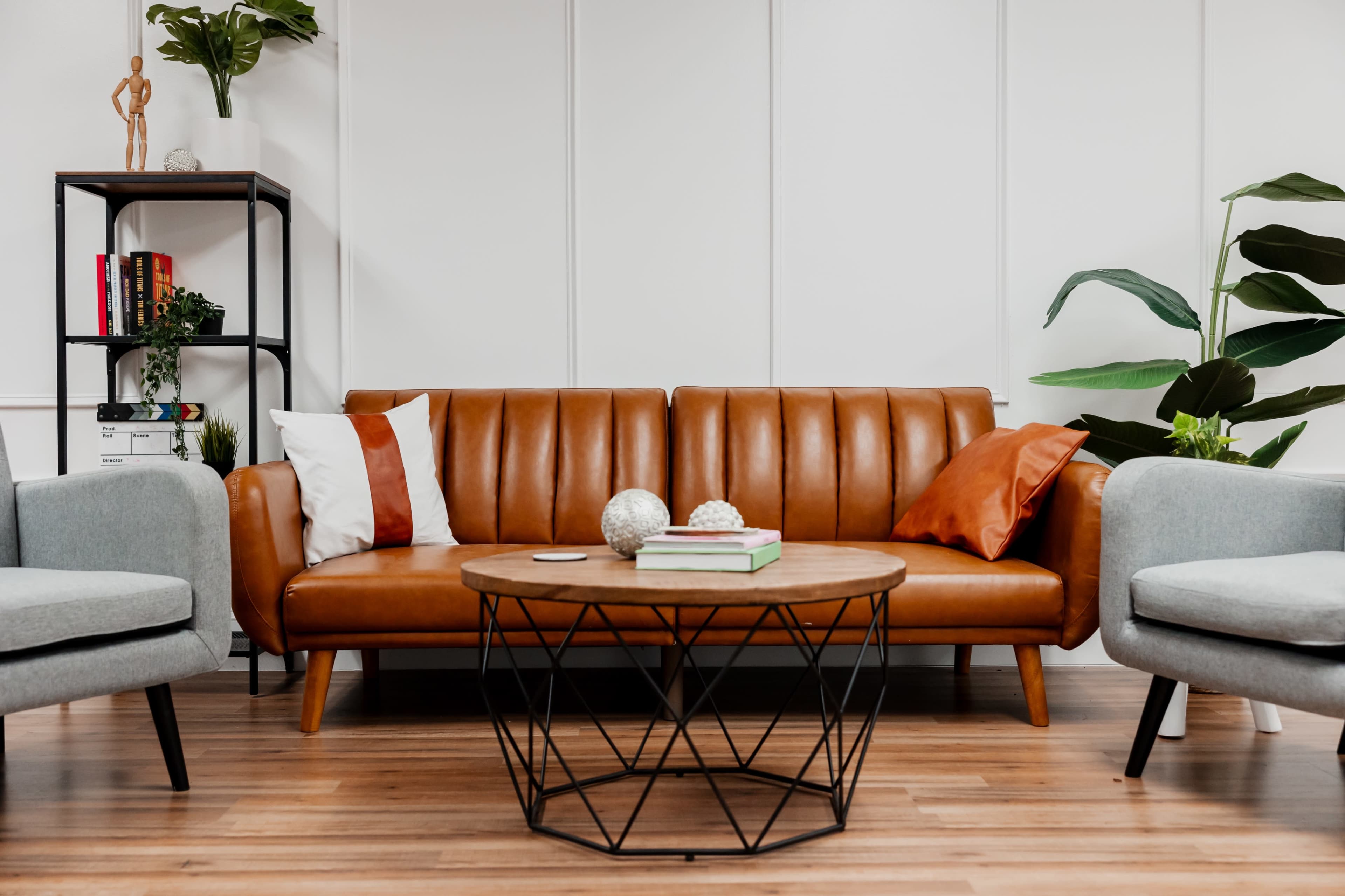 A modern living room featuring a brown leather sofa with decorative cushions, a geometric coffee table, and green plants in the background.
