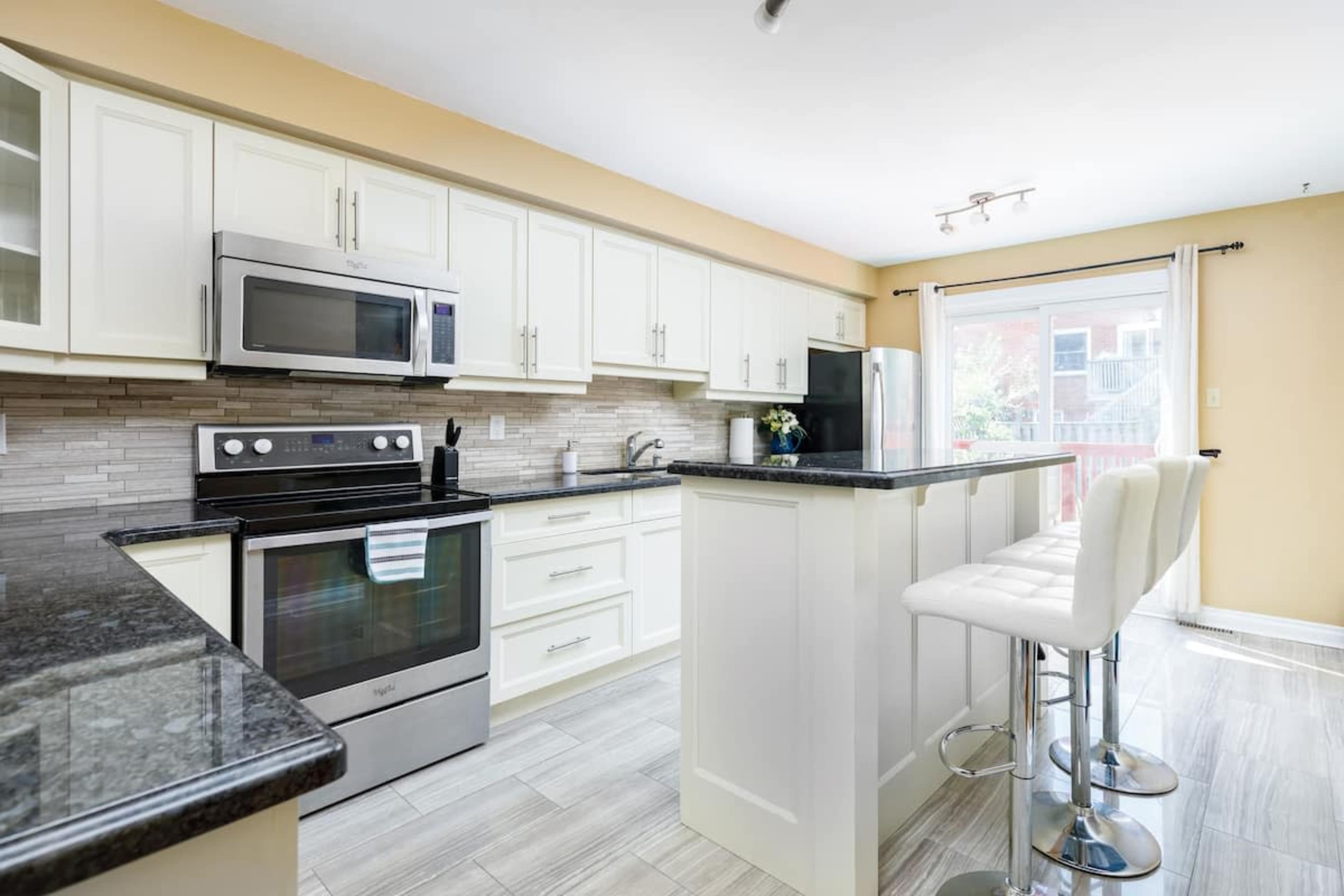 A modern kitchen features light cabinets, a dark countertop, stainless steel appliances, and a seating area with bar stools.
