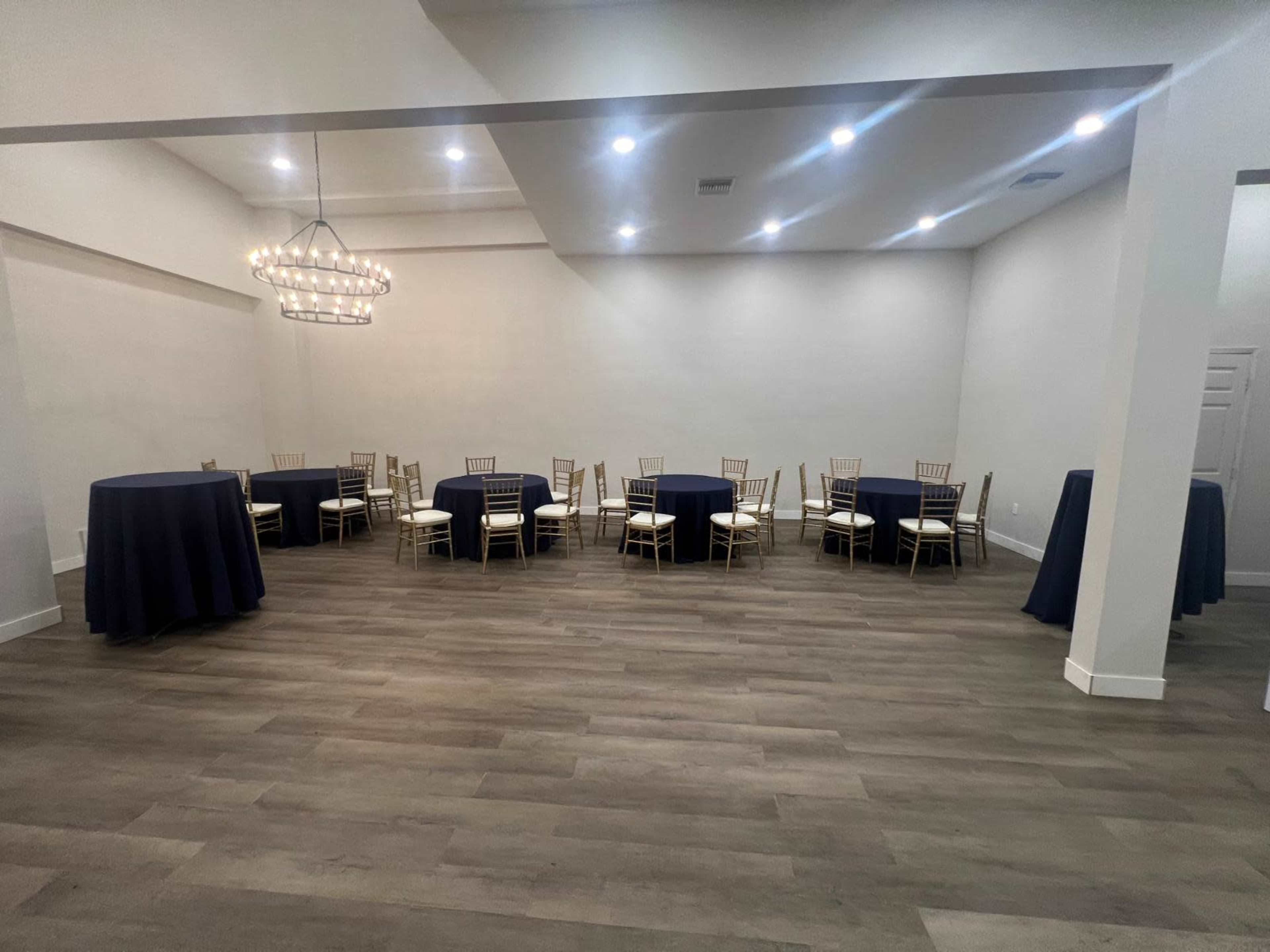The image shows a spacious event room with several round and high-top tables covered in navy tablecloths and white chairs, illuminated by a modern chandelier.