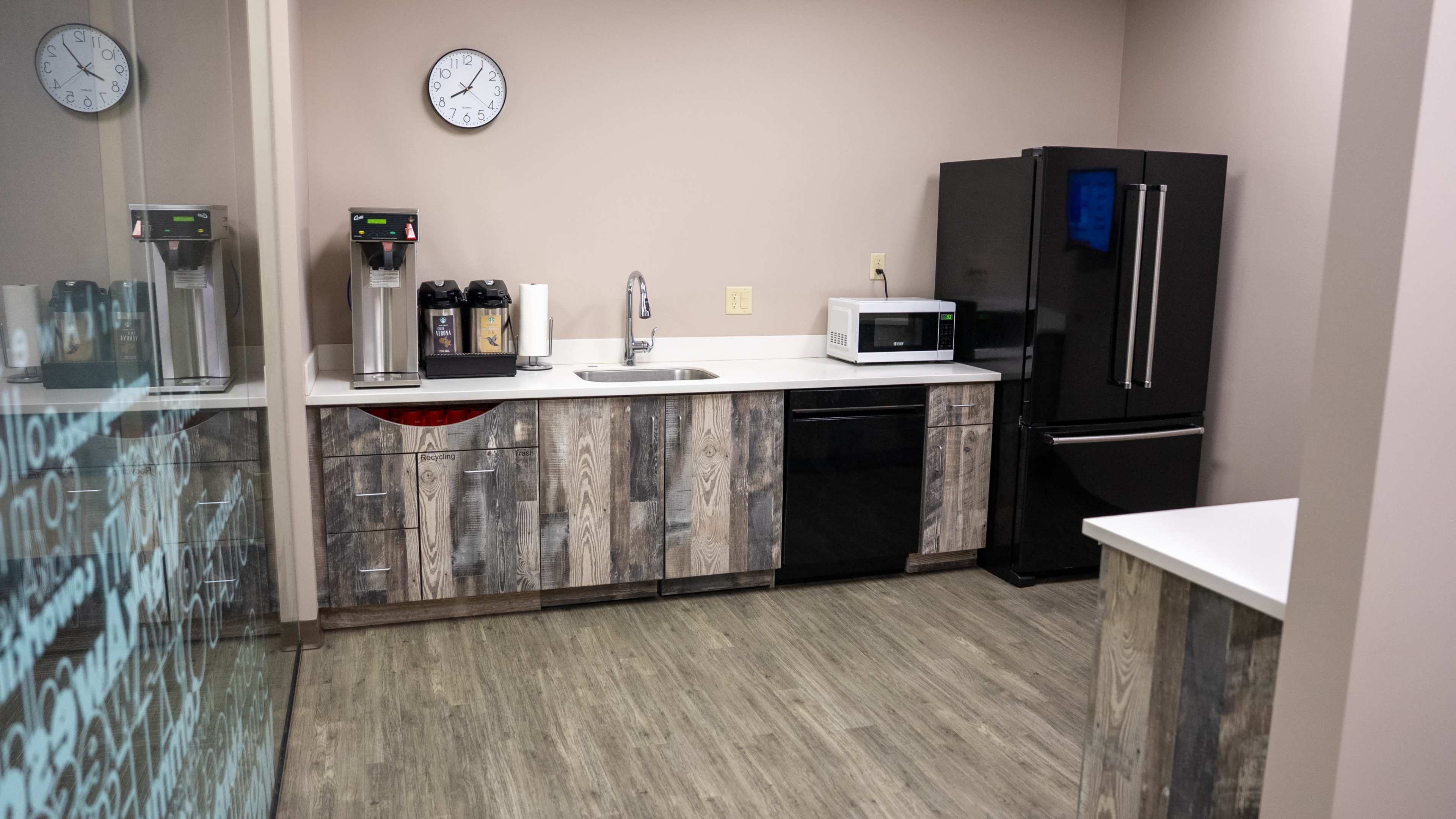 The image shows a modern kitchen area with a coffee station, a sink, a microwave, and a black refrigerator, all set against a backdrop of wood-patterned cabinetry and light-colored walls.