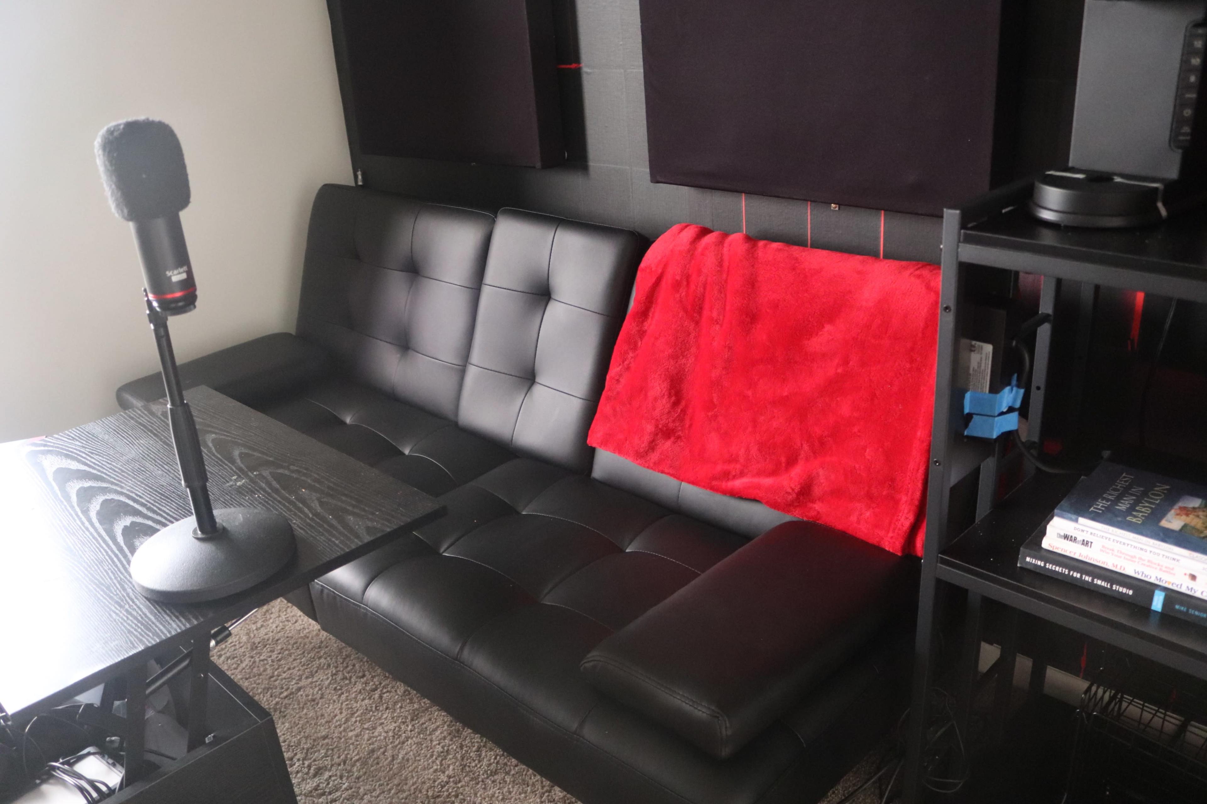 The image shows a black couch with a red blanket draped over it, beside a microphone on a stand and a wooden table in a room with soundproofing panels on the wall.