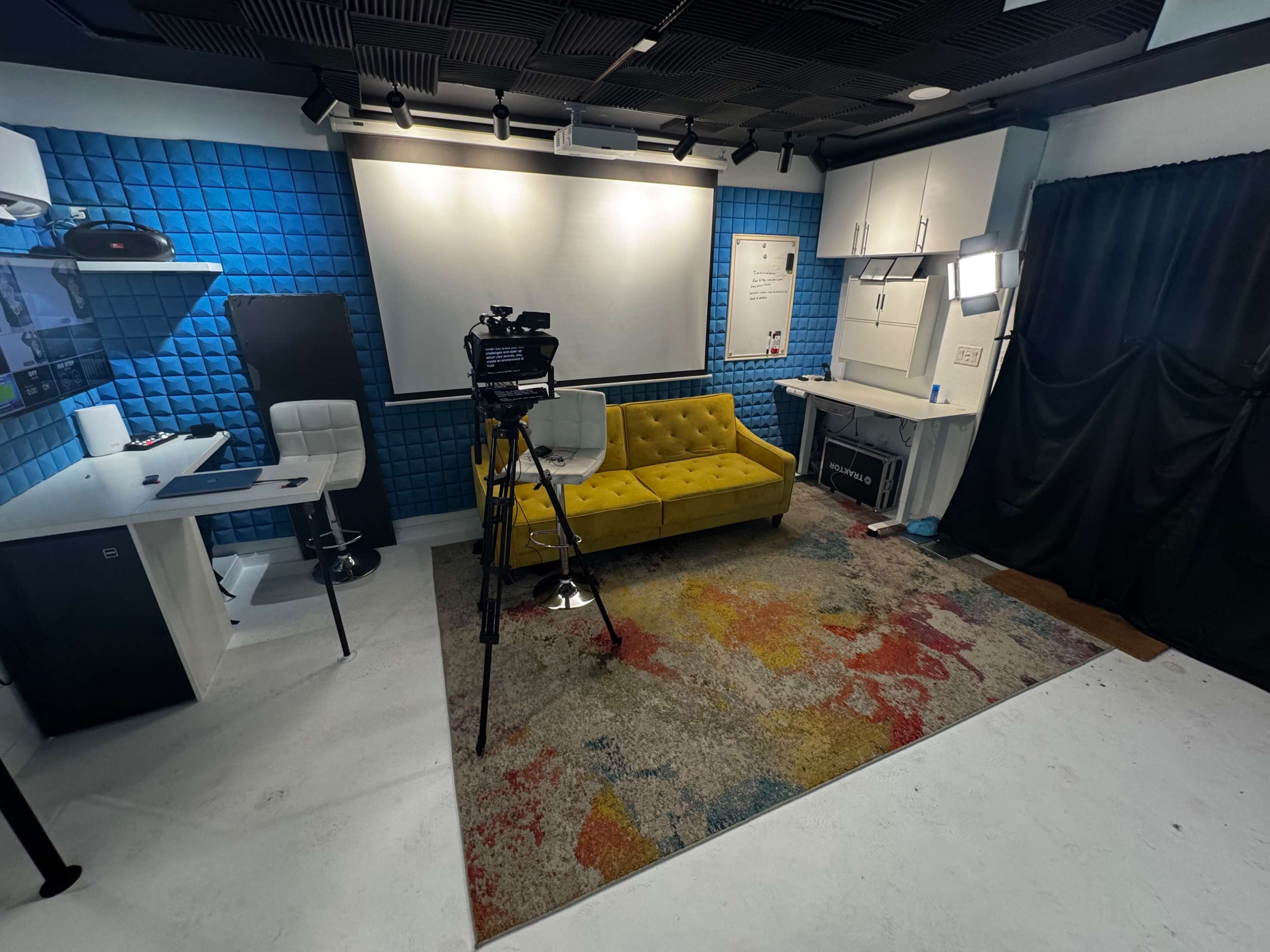 The image shows a modern studio setup with a yellow couch, a camera on a tripod, and various furniture against a blue and white backdrop.