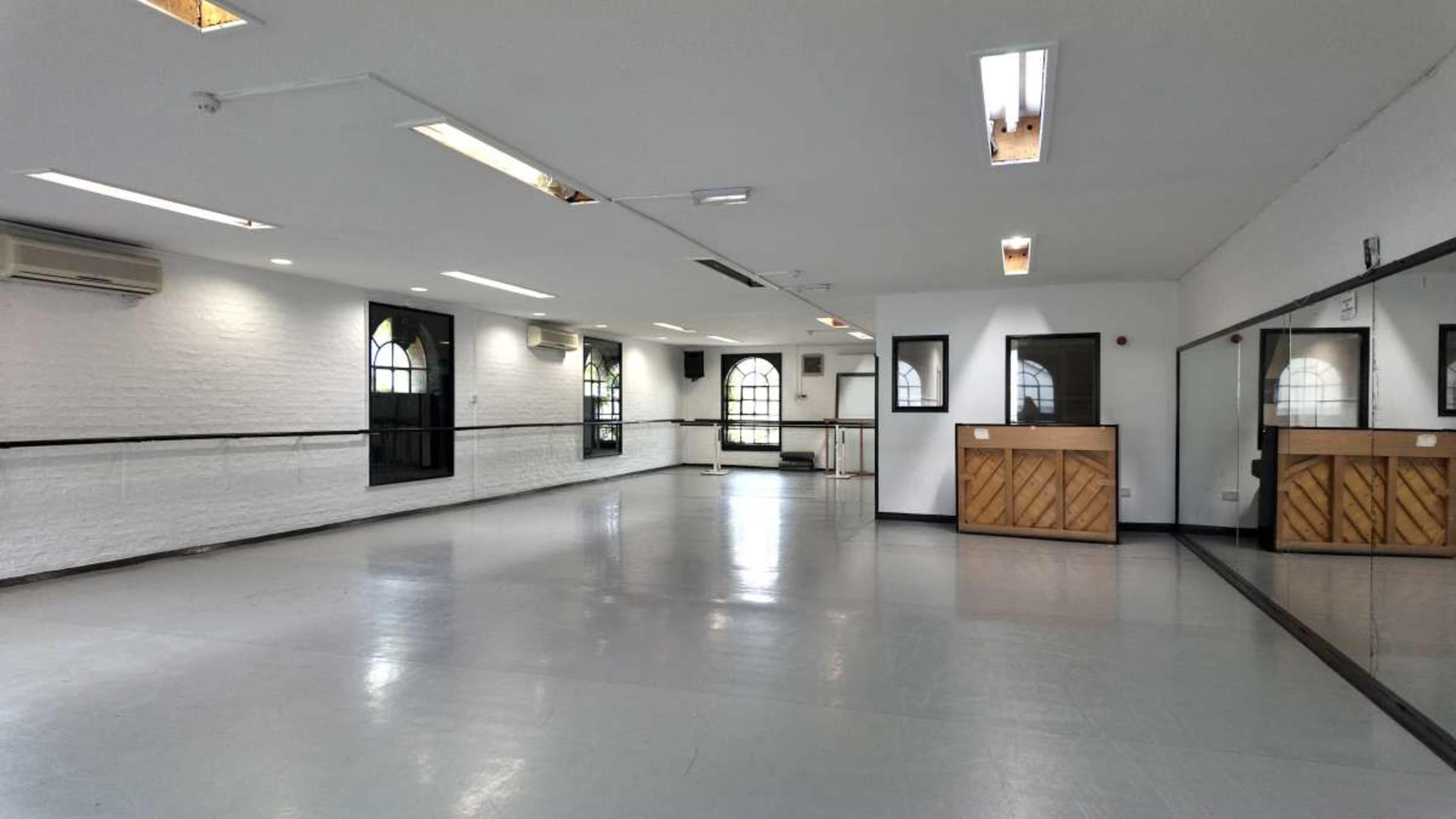 The image shows a spacious, well-lit room designed for activities such as dance or rehearsals, featuring large windows, a mirrored wall, and a wooden reception area.
