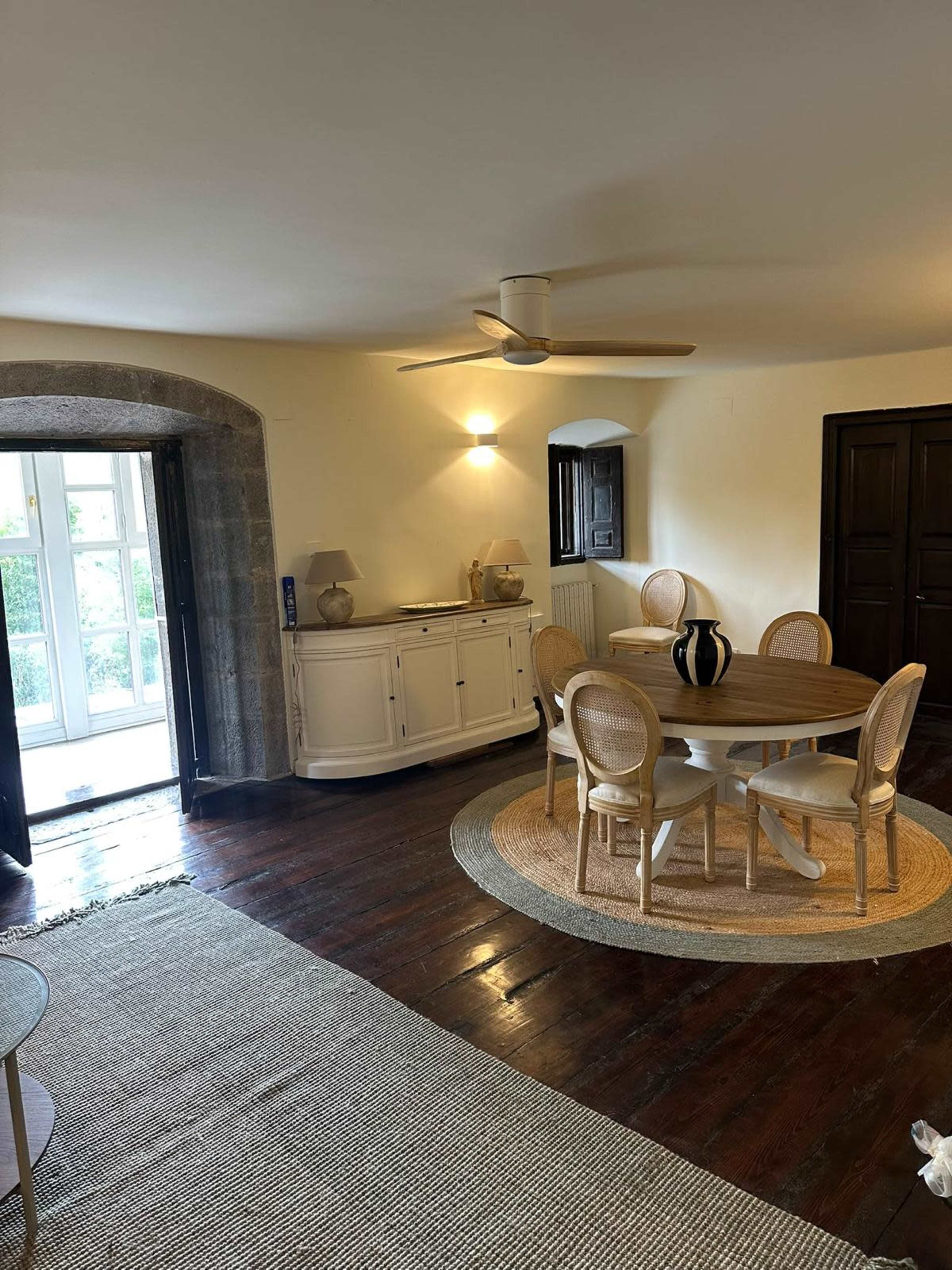 The scene features a spacious room with a round dining table surrounded by chairs, a sideboard against the wall, and large doors leading to an outdoor area.