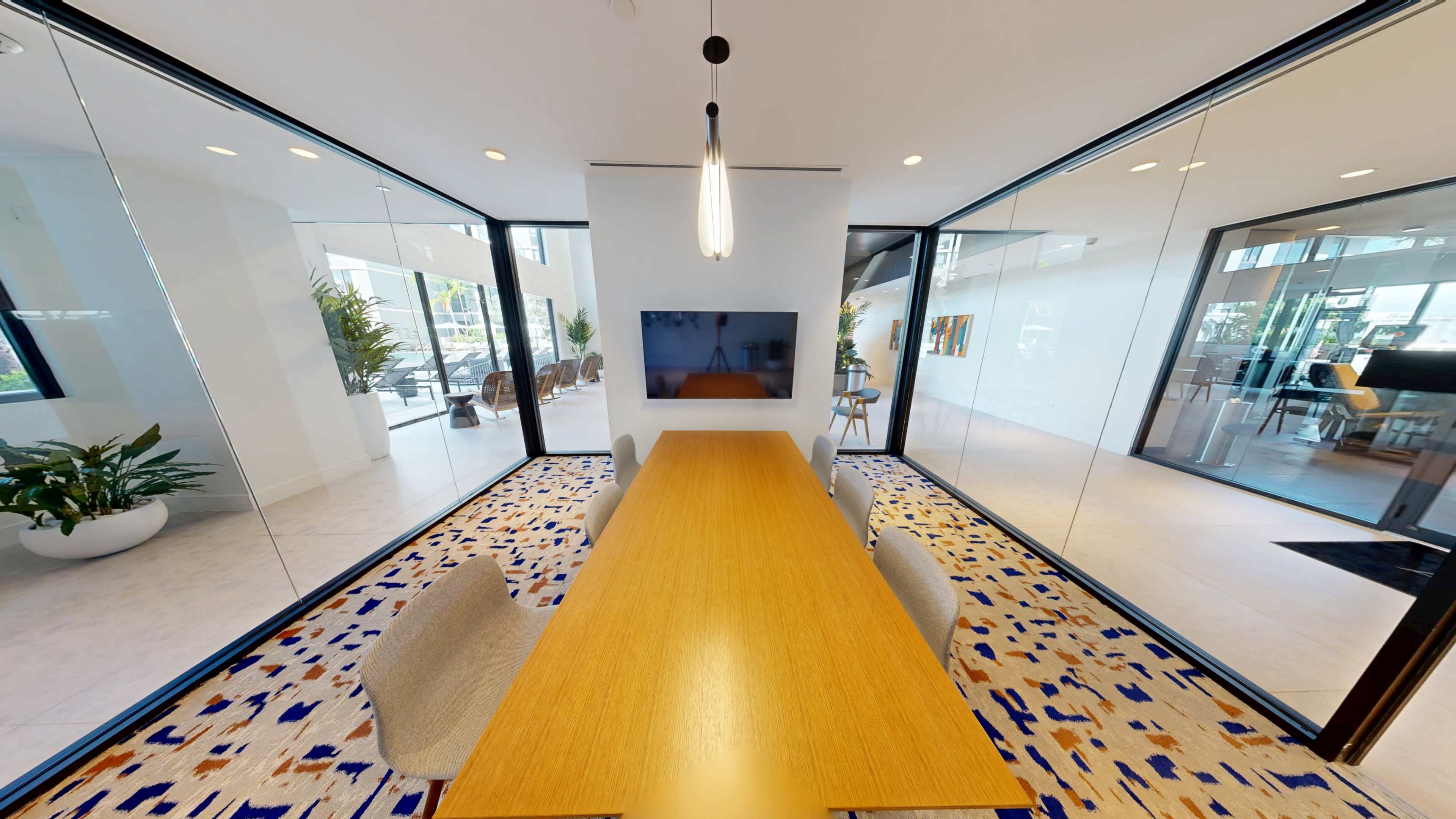 The image shows a modern conference room with a long wooden table, four chairs, and a large screen mounted on the wall, surrounded by glass walls that offer views of a brightly lit space outside.