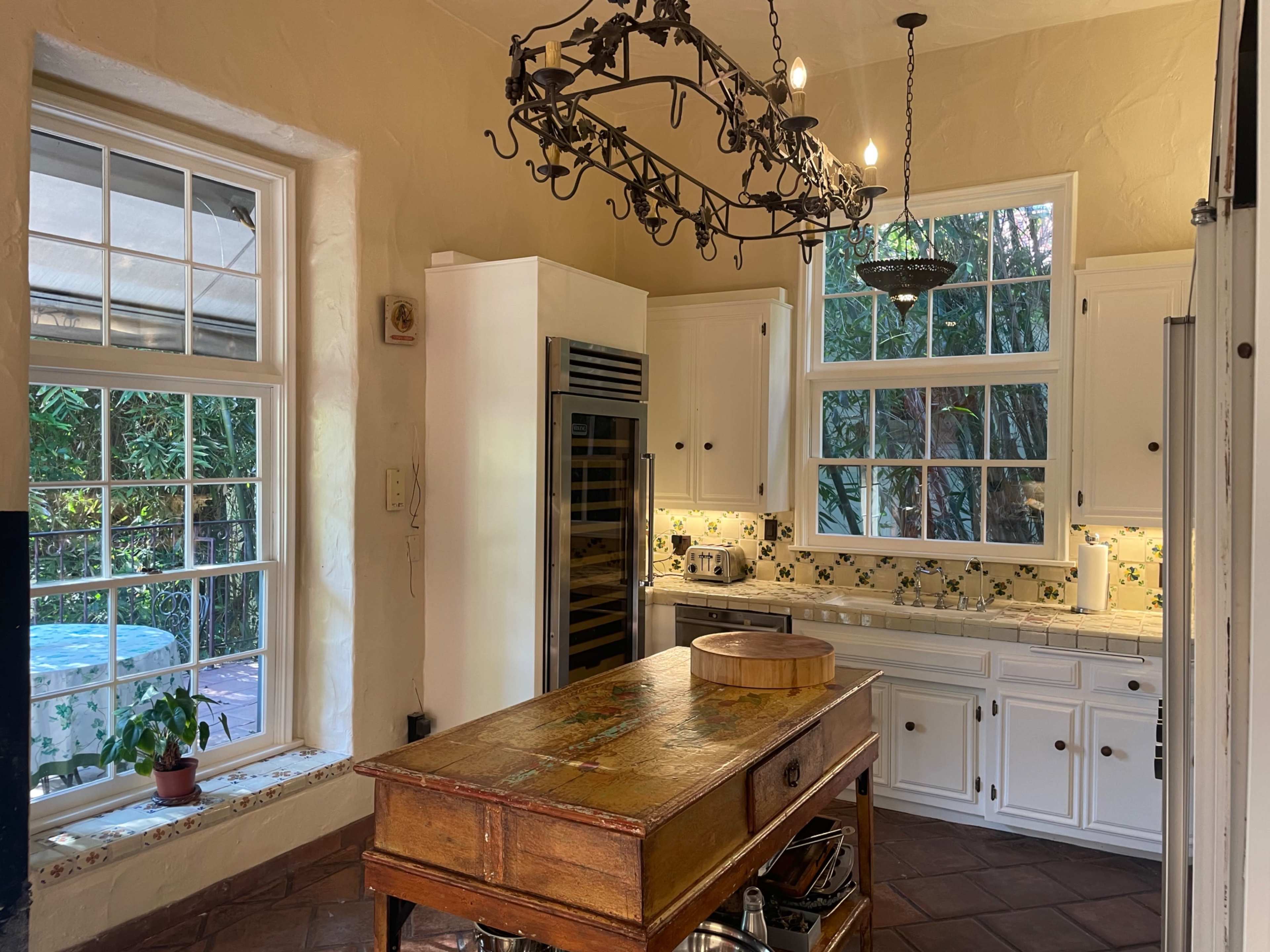 A bright kitchen with a central wooden island, white cabinetry, a decorative chandelier, and large windows overlooking greenery.