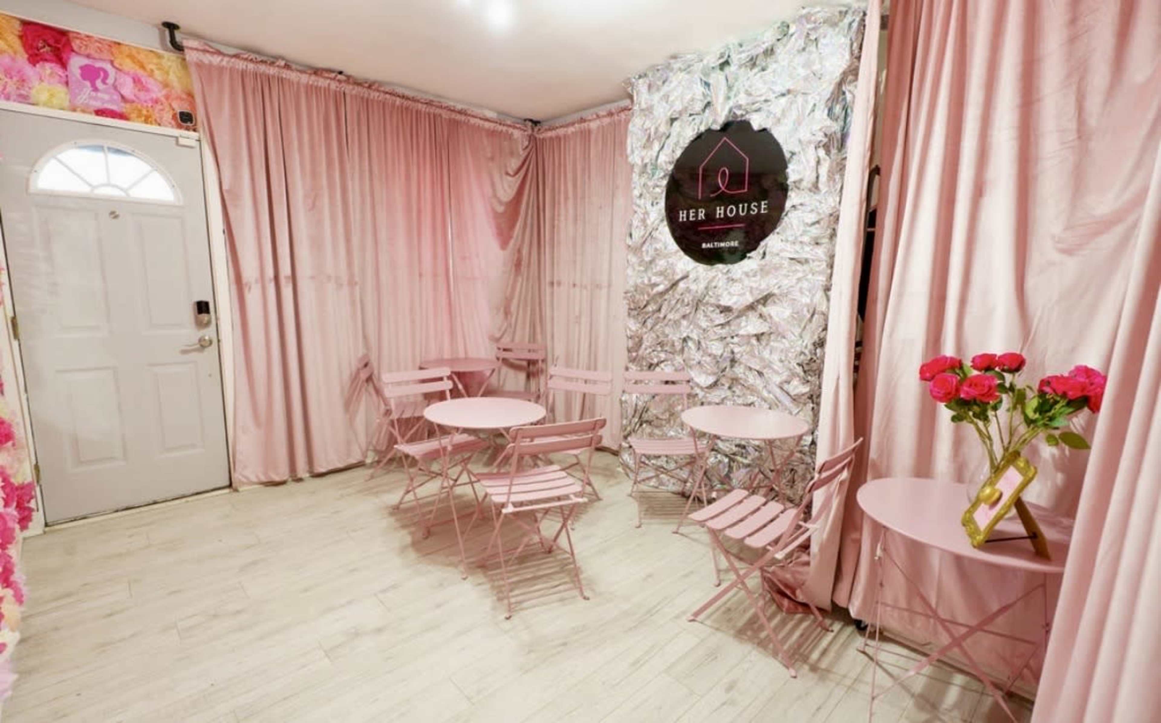 The image shows a small, stylish room with pink curtains, pink folding chairs and tables, and a shiny metallic wall with a sign reading "HER HOUSE."